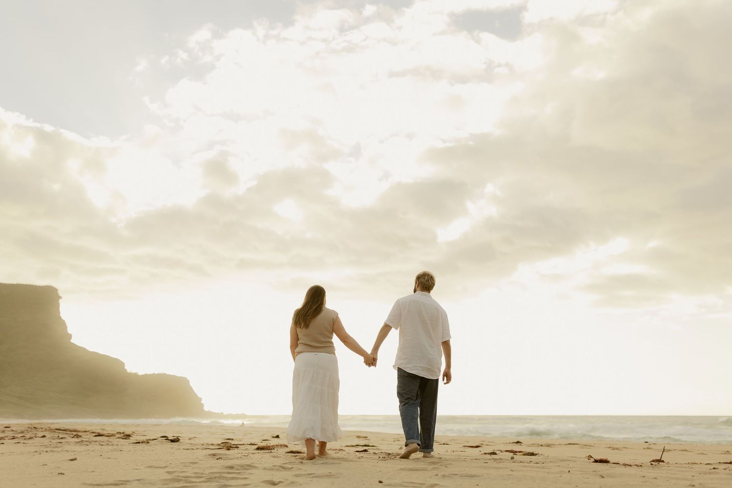 Romantic couple walking hand in hand on sandy beach during sunset with cliffs in background.