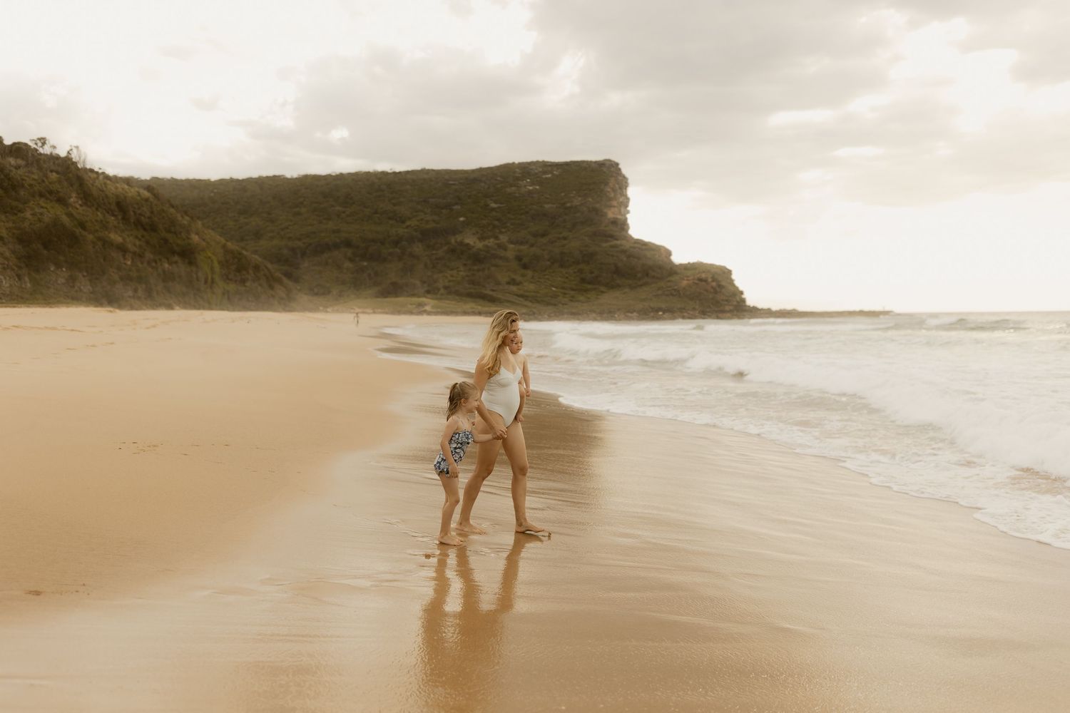Person walking along a misty beach shoreline with dramatic cliffs in the background during golden hour.