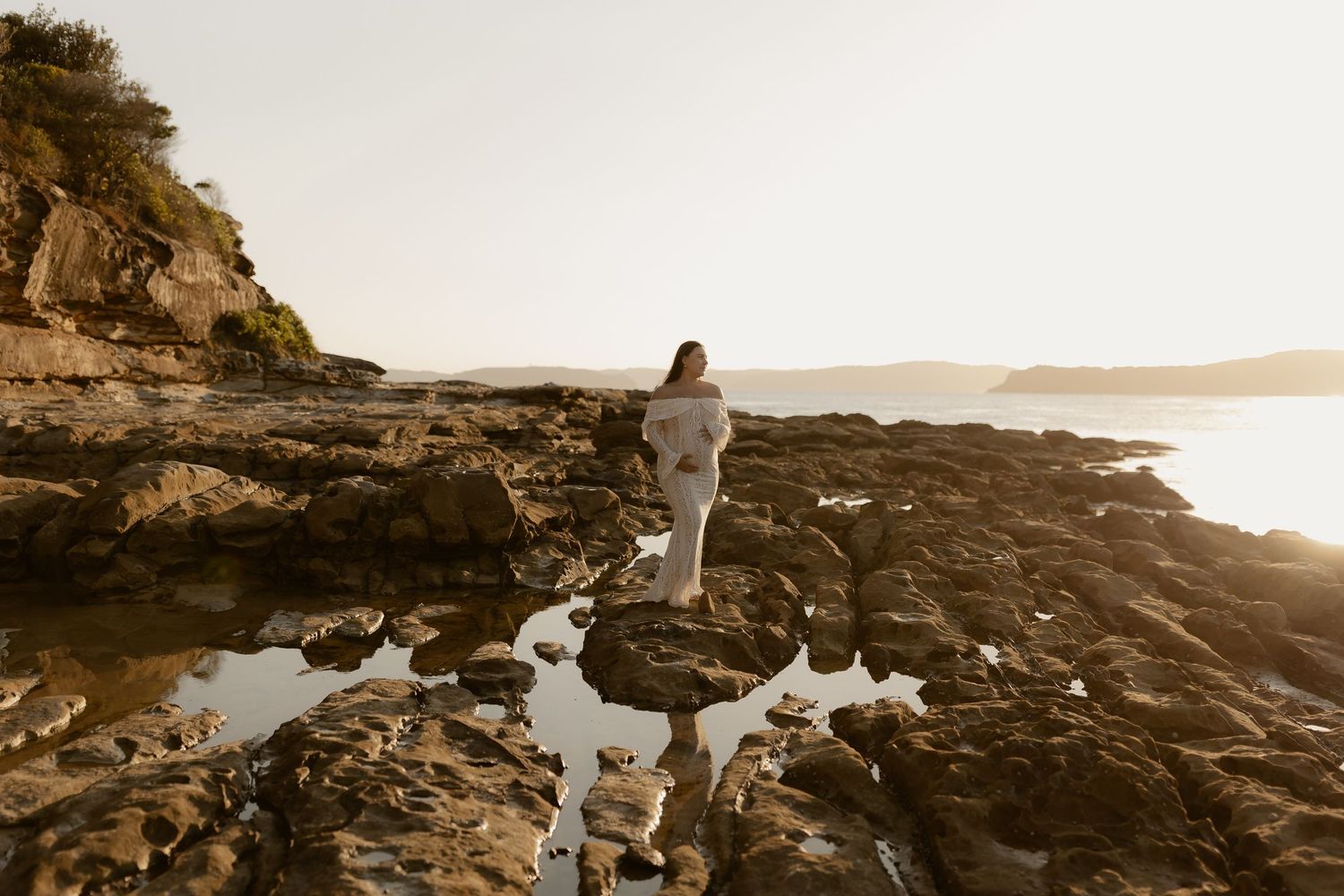 Couple standing on rocky coastal cliffs during golden sunset with ocean views.