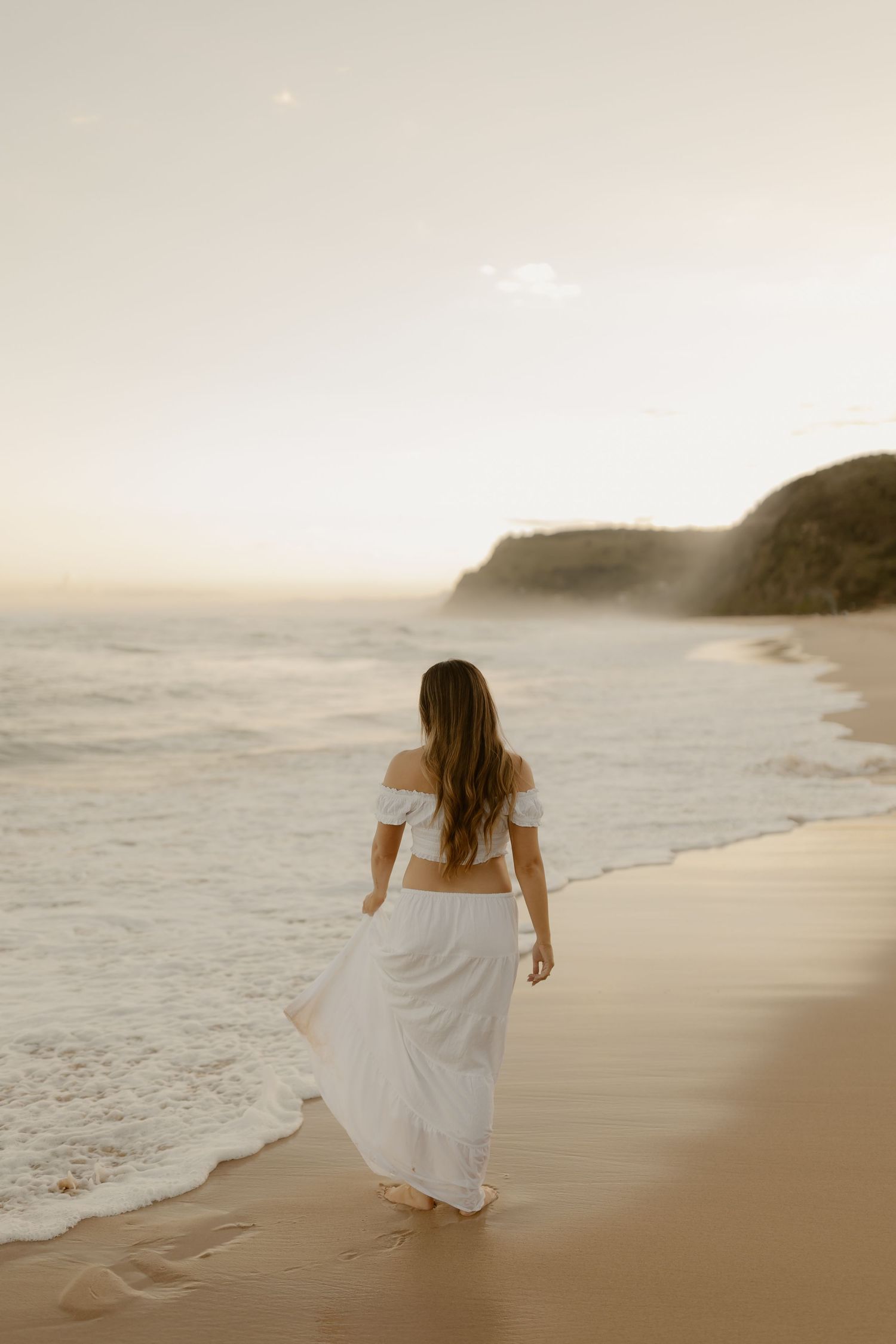 Woman in flowing white dress walks along a scenic beach at sunset with cliffs in the distance.