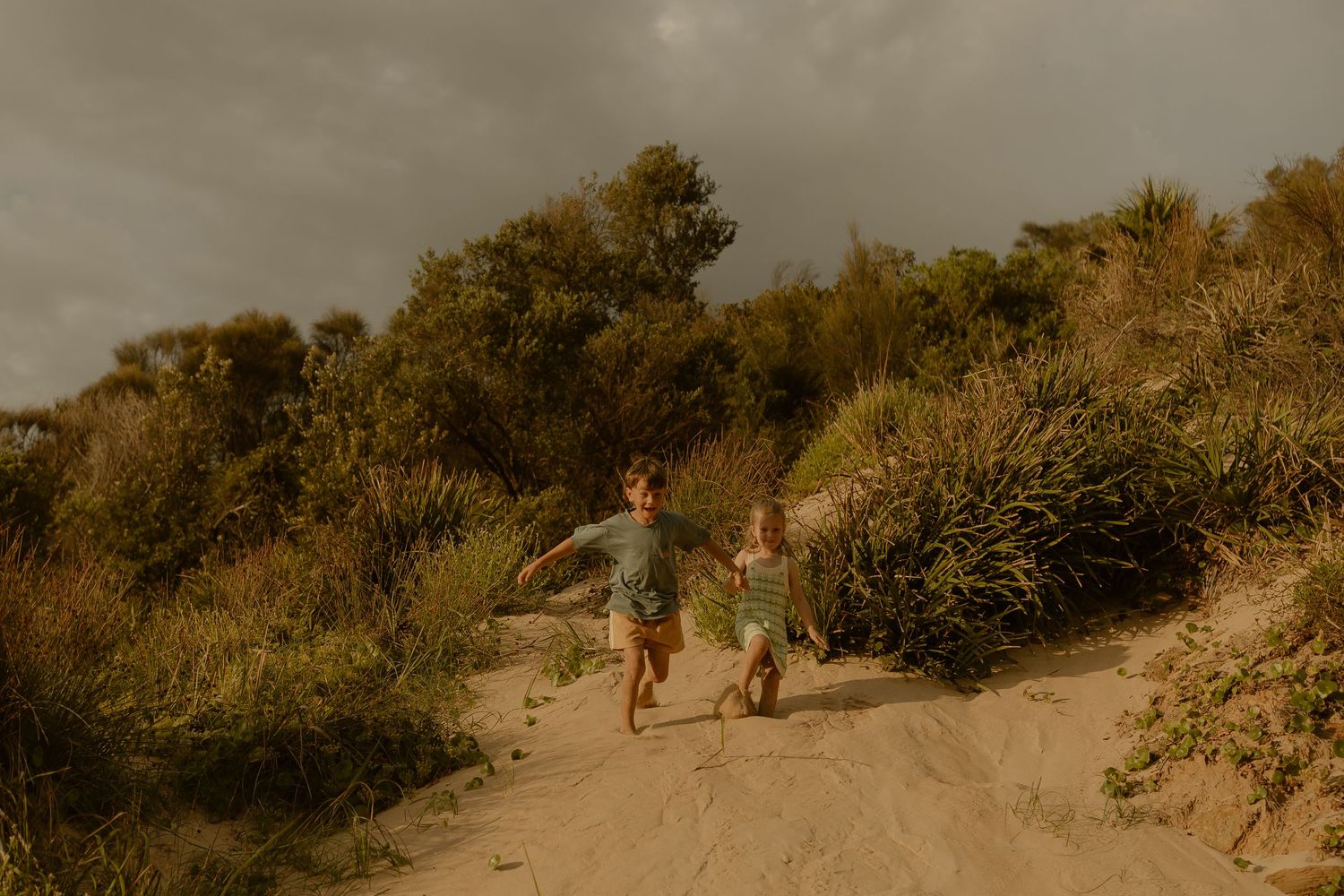 Two figures walking hand in hand up a sandy coastal path through wild grasses.
