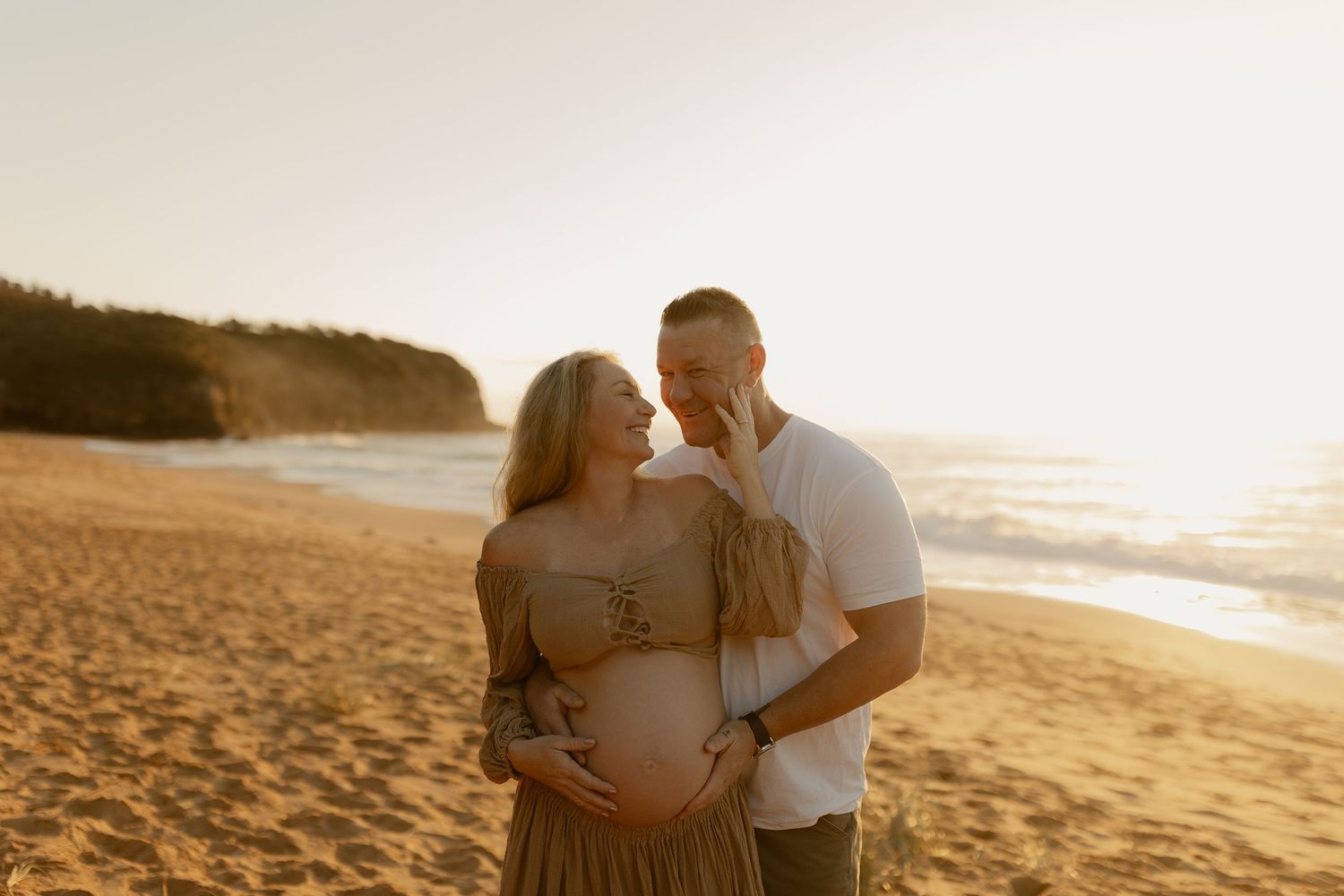Expecting couple sharing a tender moment on a sunlit beach, woman in olive dress holding baby bump.