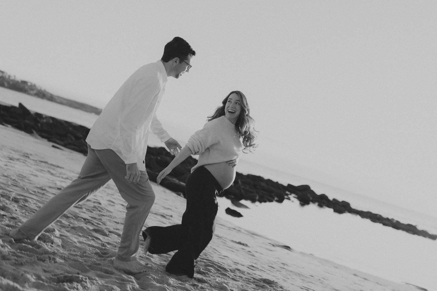 Black and white photo of a laughing pregnant couple playing on a beach near rocky shores.