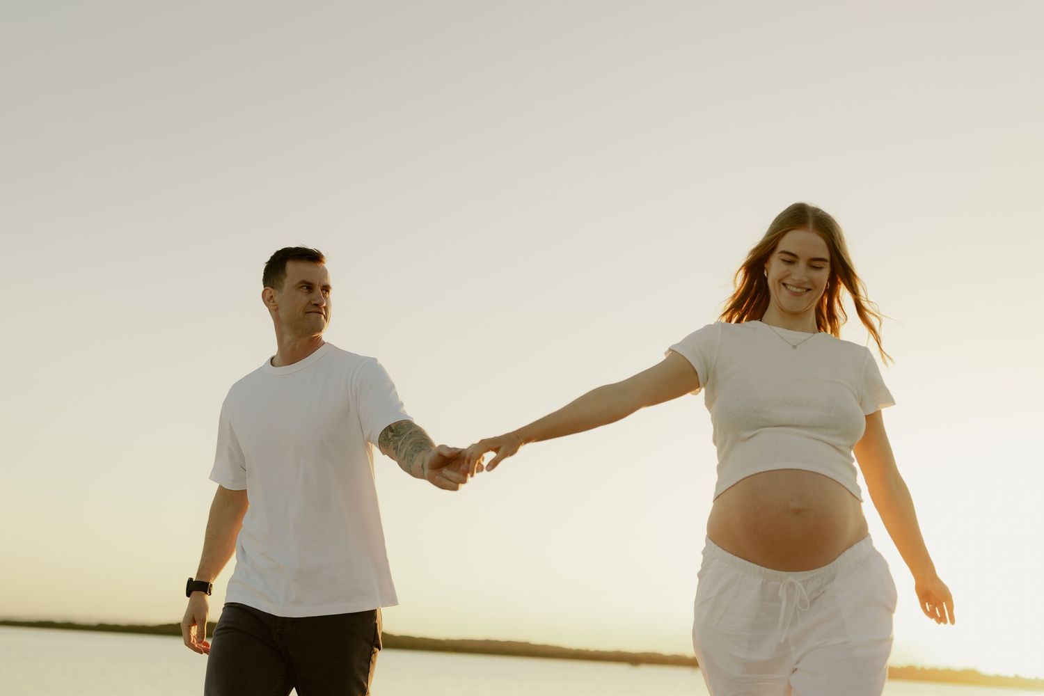 Pregnant couple holding hands on a beach at sunset, man in white t-shirt, woman in white crop top showing baby bump.