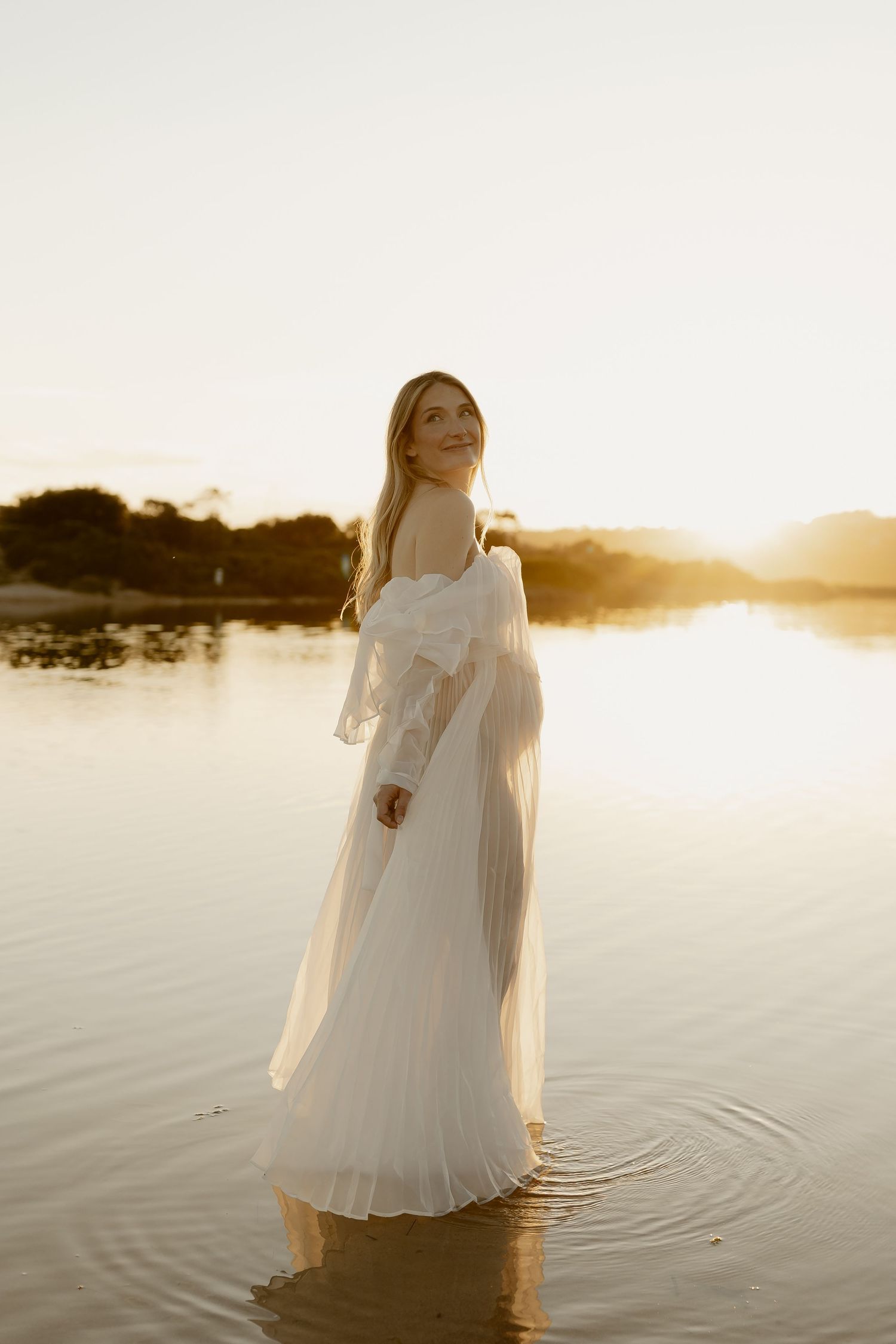 A person in a flowing white dress stands in calm lake waters at sunset creating a dreamy ethereal scene.