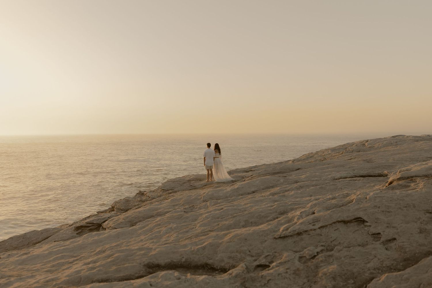 Two figures stand on rocky coastal cliffs overlooking a misty ocean at sunset.
