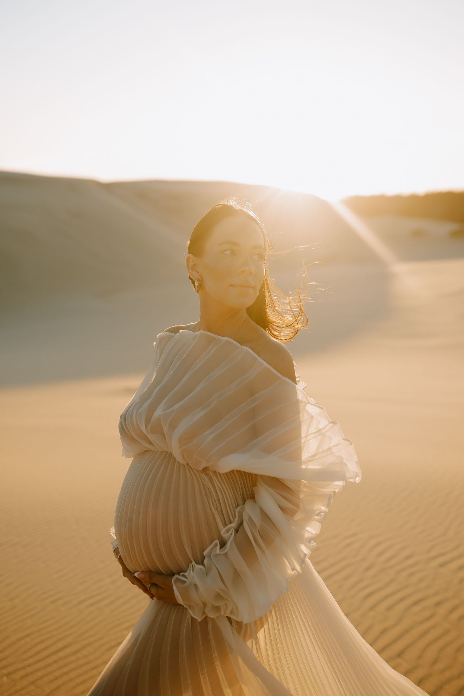 A figure in flowing white fabric sits on desert sand dunes at golden hour with sunlight glowing behind them.