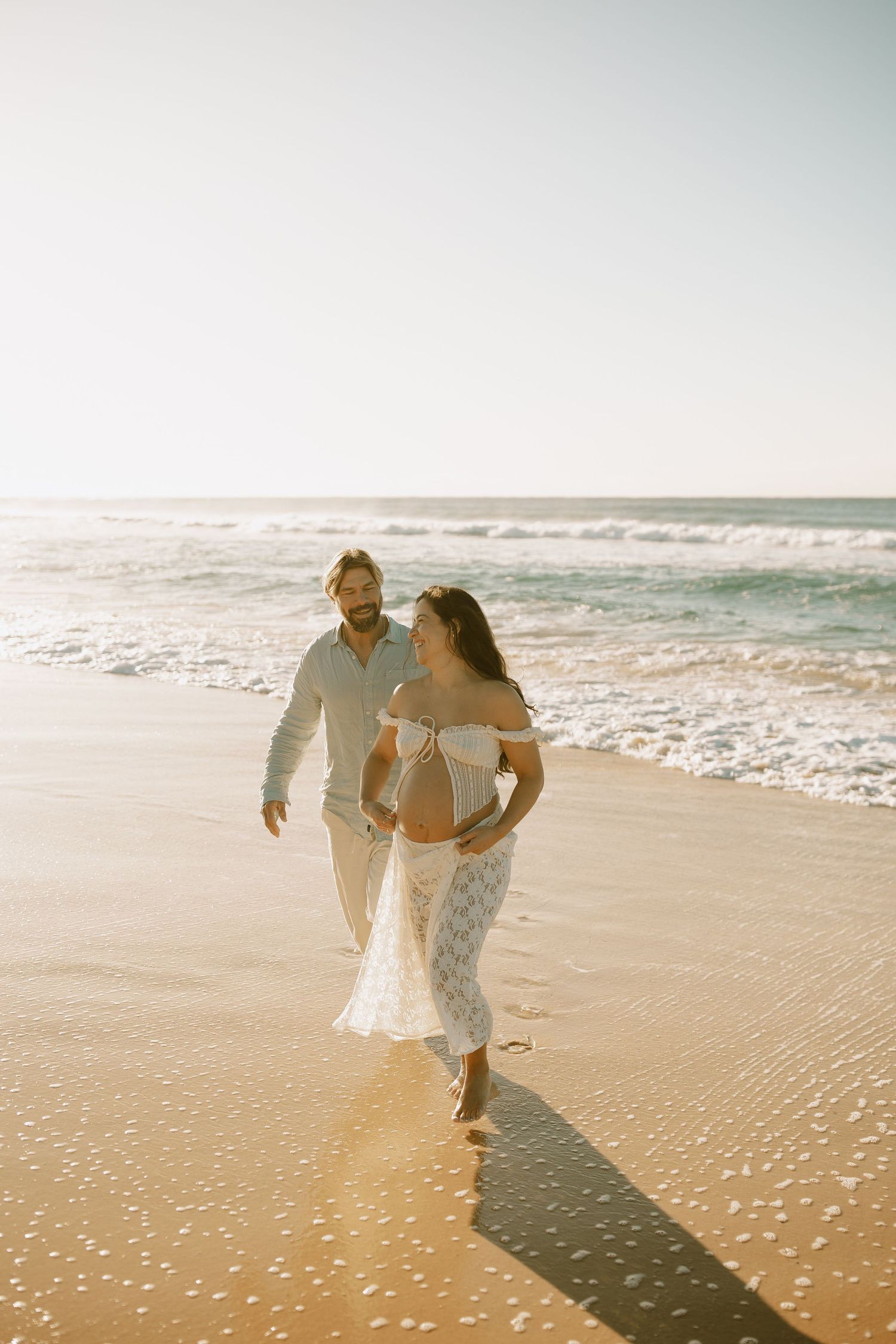 Couple walks along sandy beach shoreline during sunset maternity photo session.