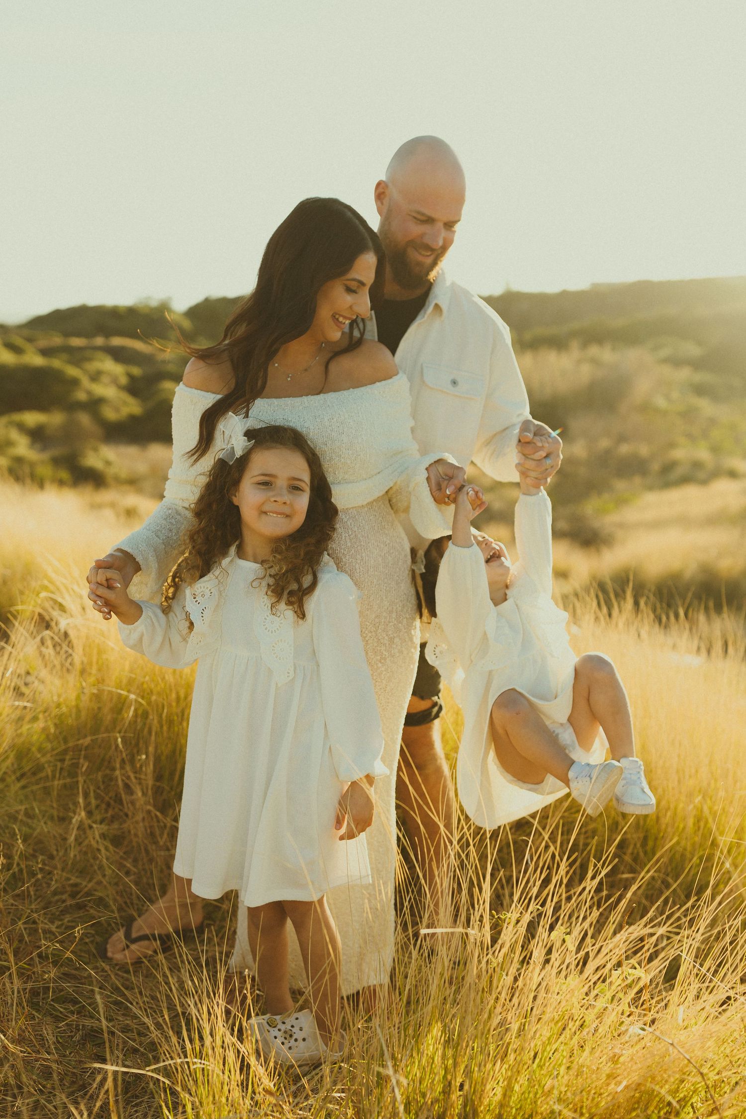 A family in white clothing embraces while standing in a sun-drenched meadow of tall grass.