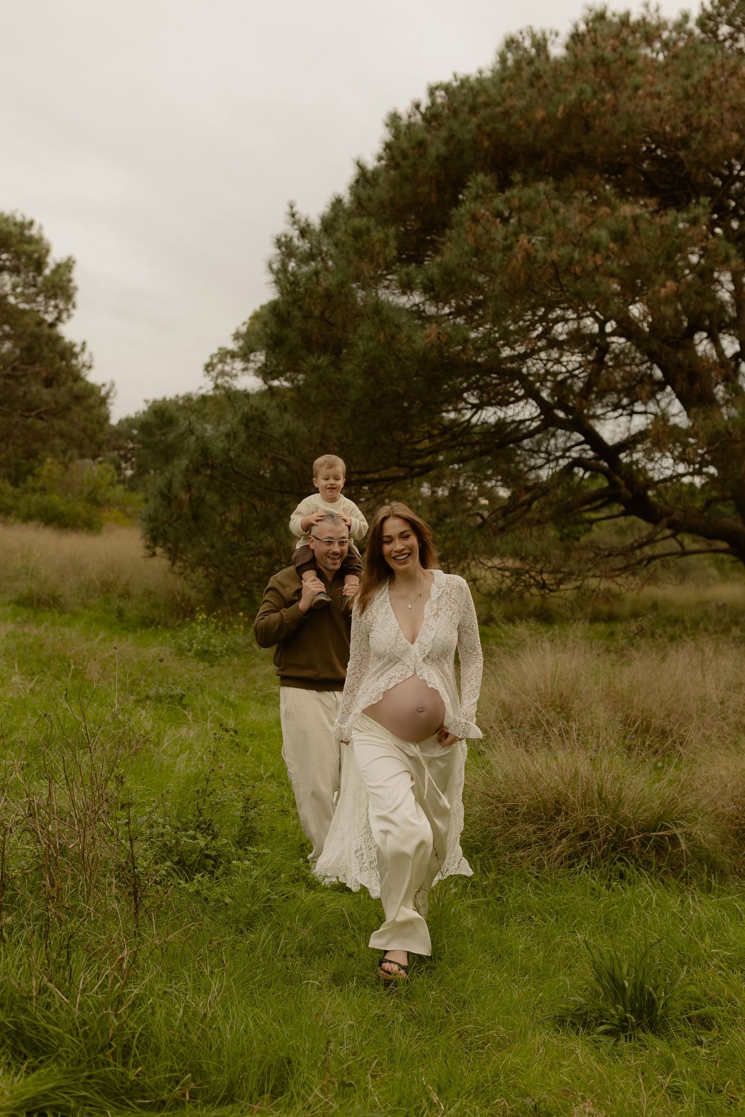 Pregnant woman in white lace dress walks with partner carrying toddler on shoulders through green field.