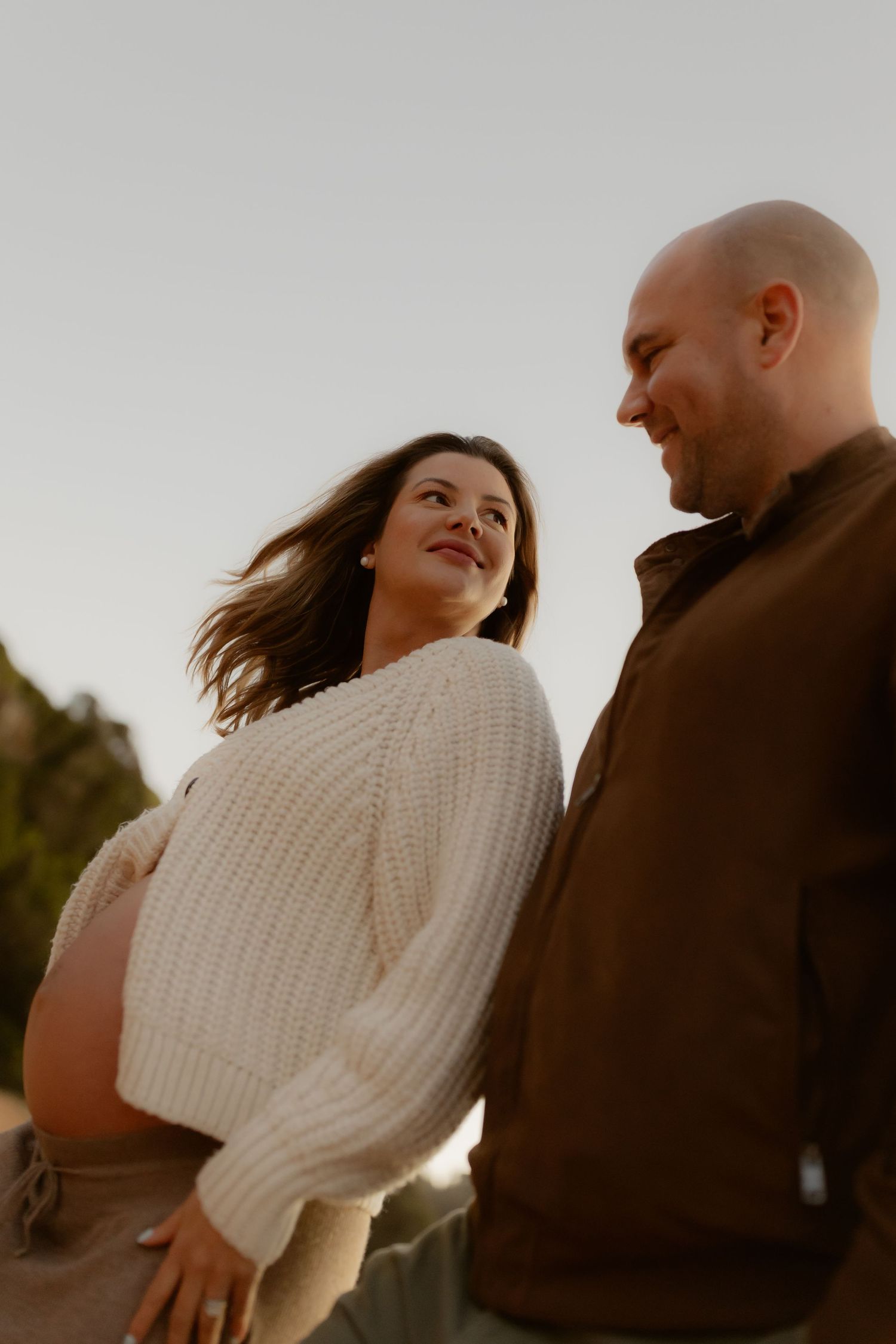 Smiling pregnant woman in cream knit sweater standing with bald partner outdoors at golden hour.