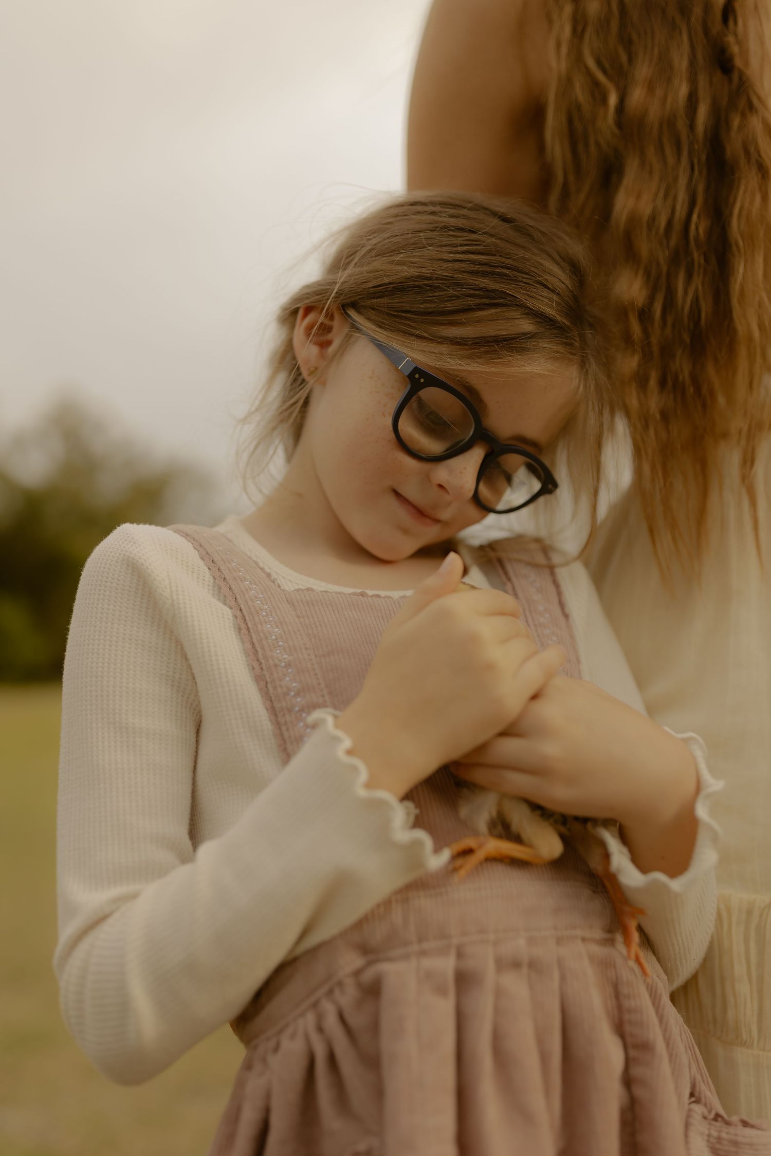 A young girl with black glasses holds a small chick while standing outdoors in a field at golden hour.