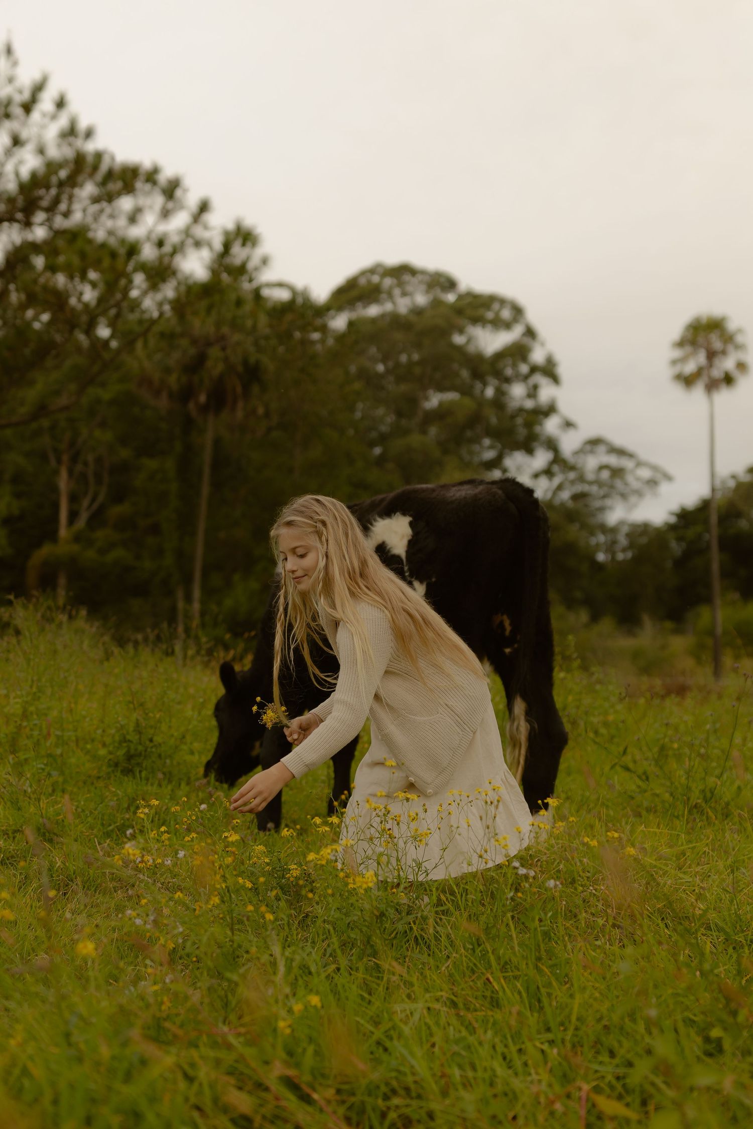 Blonde woman in white dress kneeling beside a black and white calf in a wildflower meadow.
