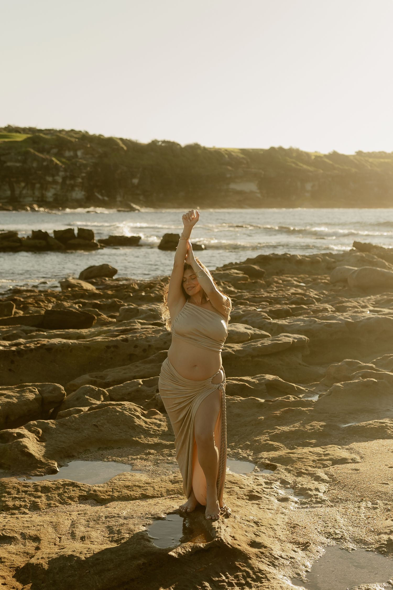 Pregnant woman in beige dress poses on rocky beach at golden sunset with ocean waves and cliffs in background.
