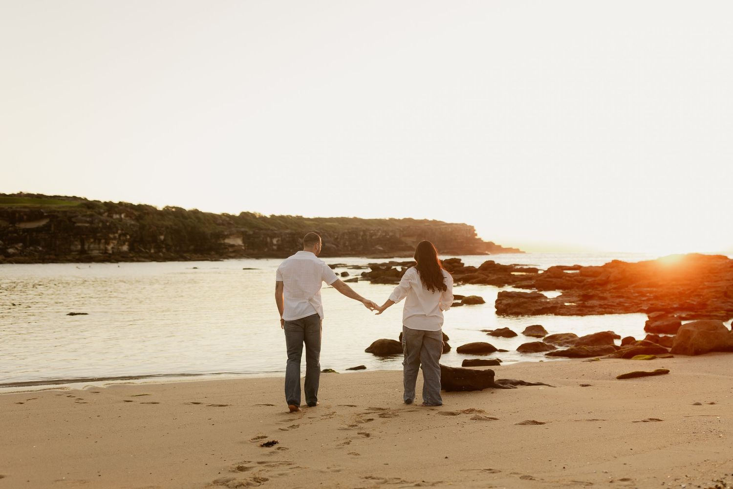Couple holding hands facing the ocean on a sandy beach during a warm golden sunset.
