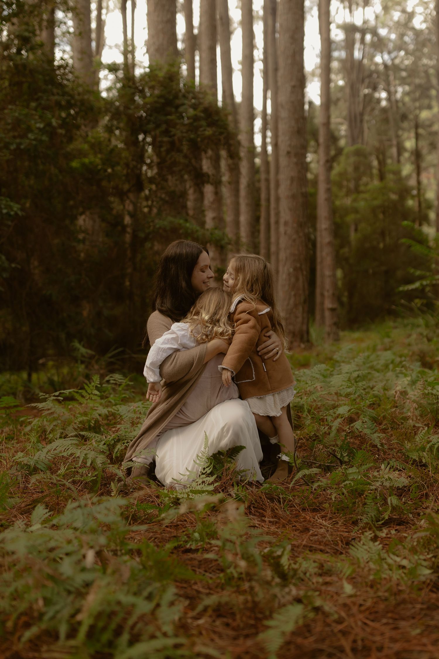 Mother kneeling in forest embracing two young children among ferns and tall pine trees.