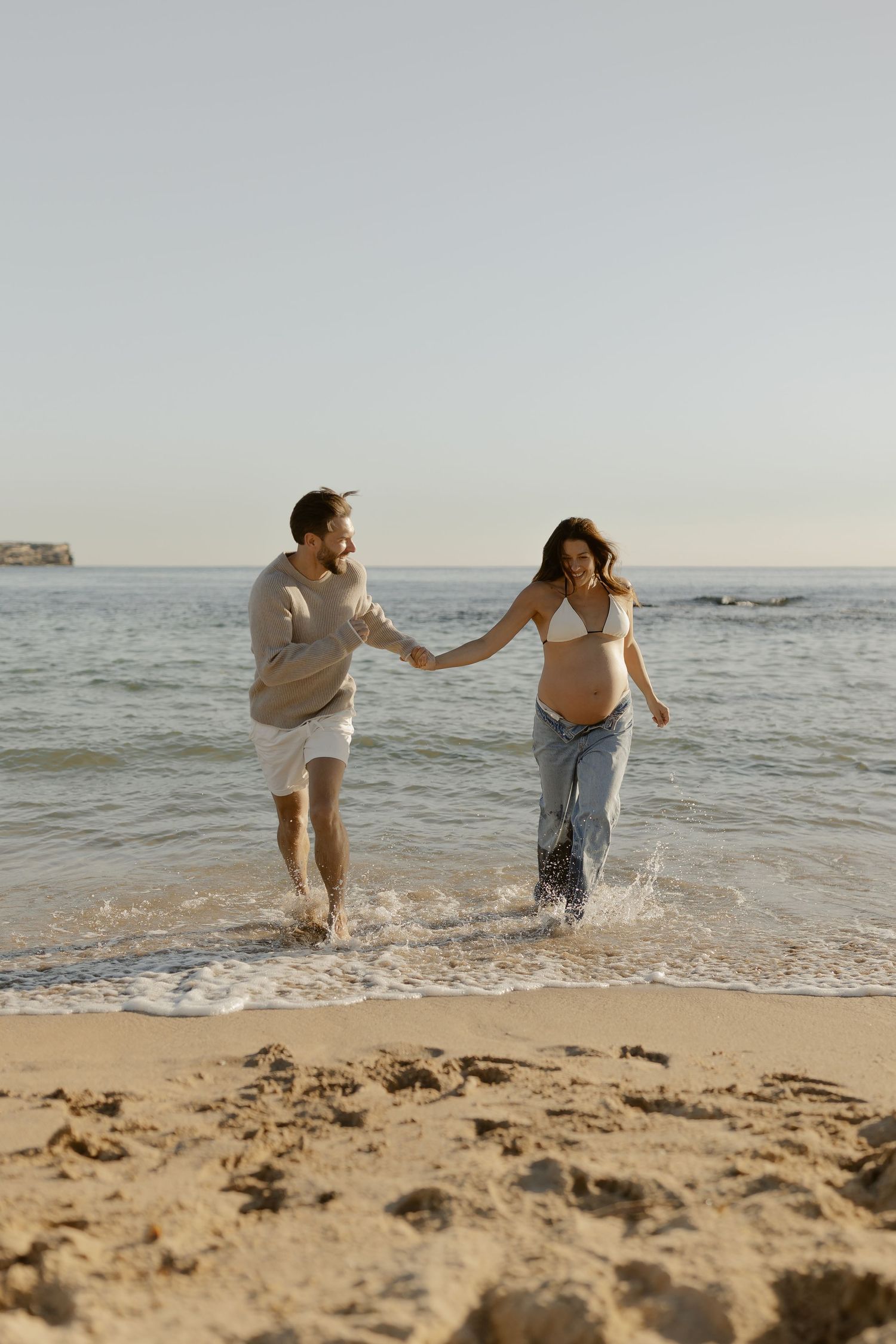 Couple runs through shallow ocean water during sunset beach photoshoot.