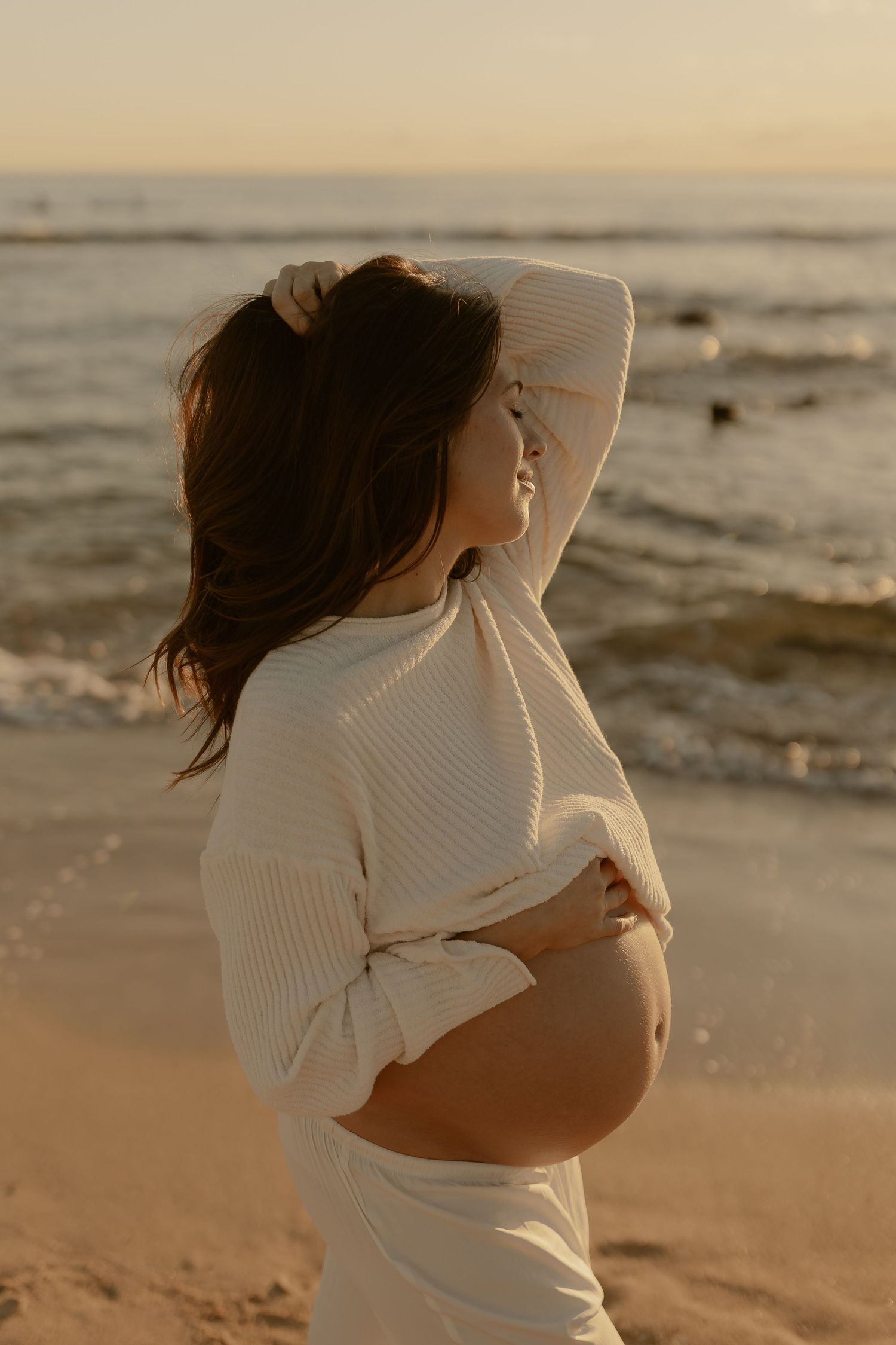 A pregnant woman in a white sweater poses on the beach during golden hour.
