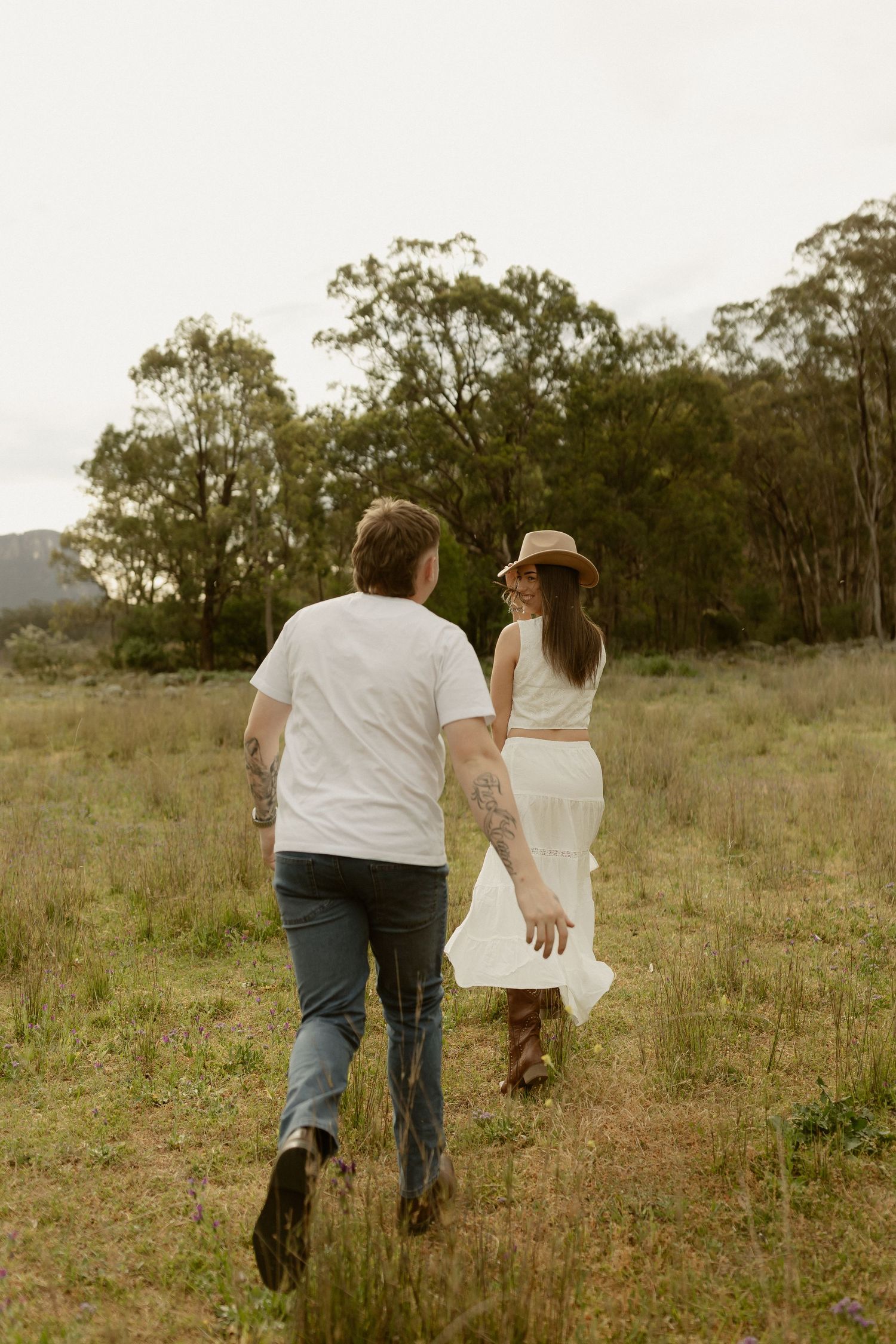 A couple walking hand in hand through a grassy field with eucalyptus trees in the background at sunset.