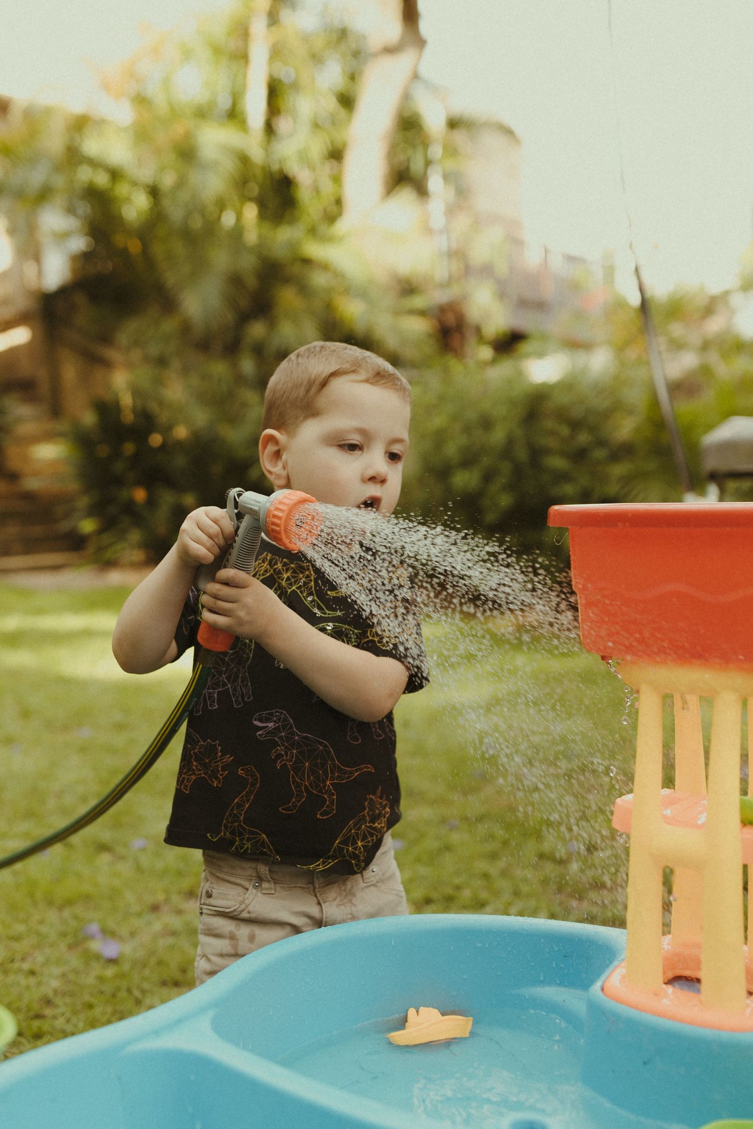 A child sprays water from a garden hose at a blue water table in the backyard.