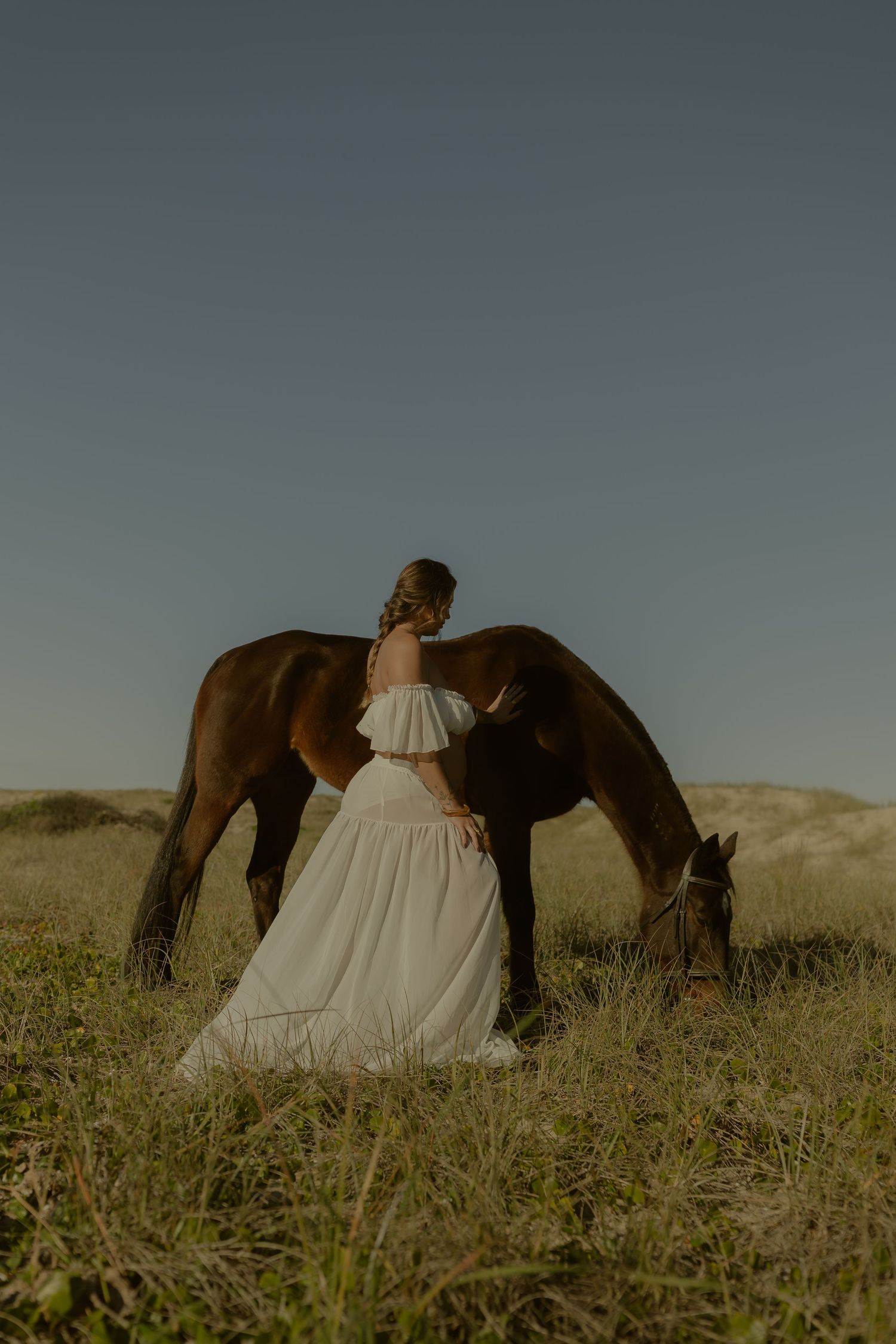Woman in white off-shoulder gown stands beside a grazing brown horse in a golden field at dusk.
