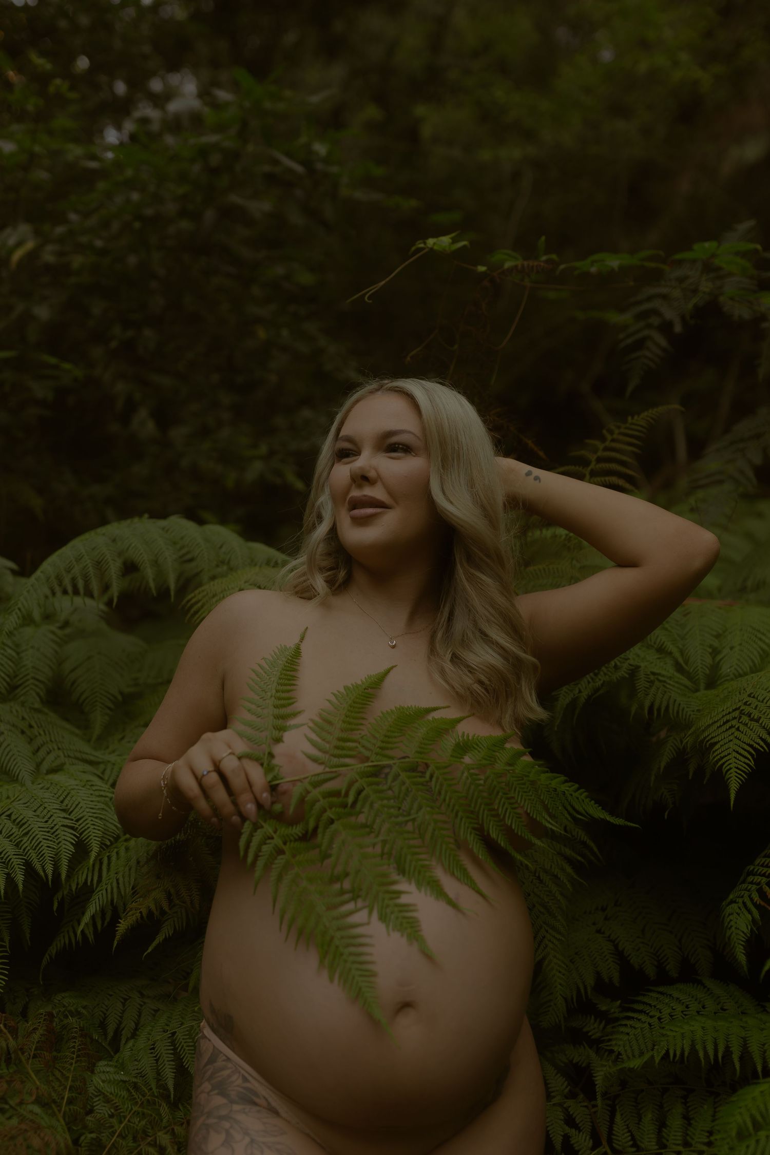 Pregnant woman posing outdoors among lush green ferns, holding a fern frond, smiling in a natural forest setting.