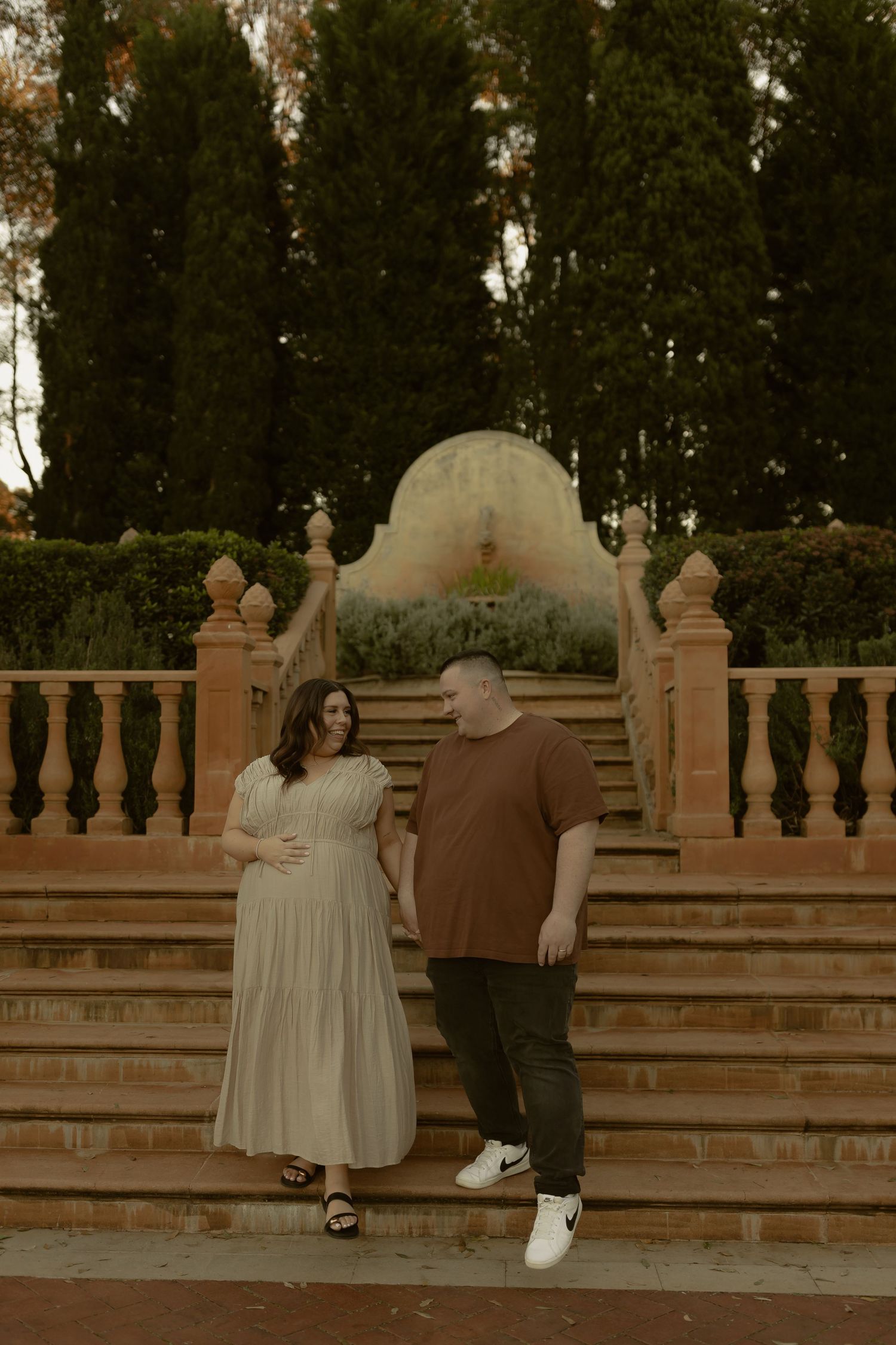 Couple poses on ornate stone steps in a garden, woman in cream dress cradling baby bump, man in brown tee.