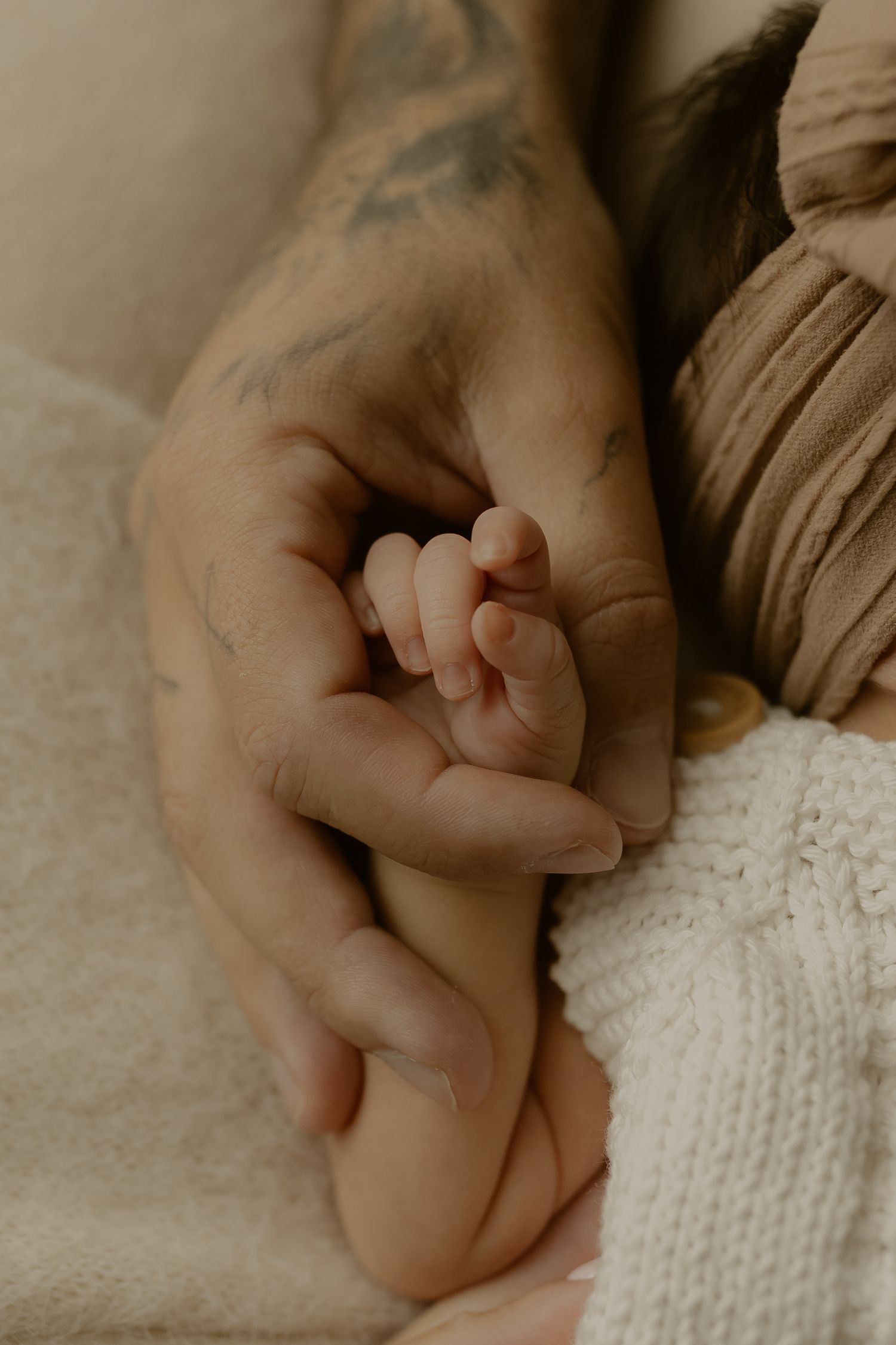 A tattooed adult hand gently holds a tiny newborn baby's hand, symbolizing parental love and new life.