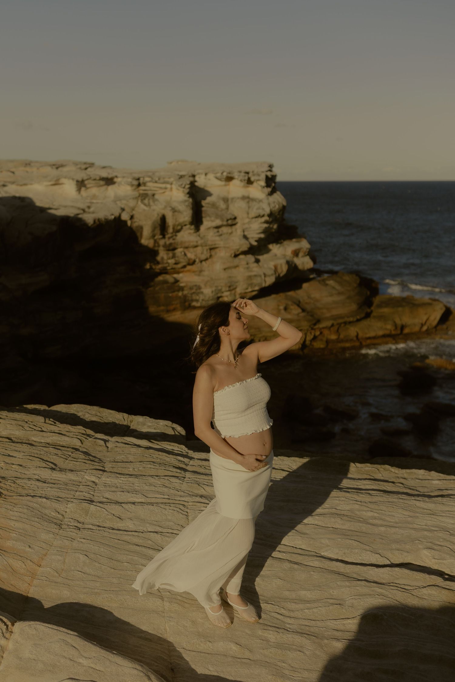 Pregnant woman in white strapless dress standing on coastal rocks at golden hour with ocean views.