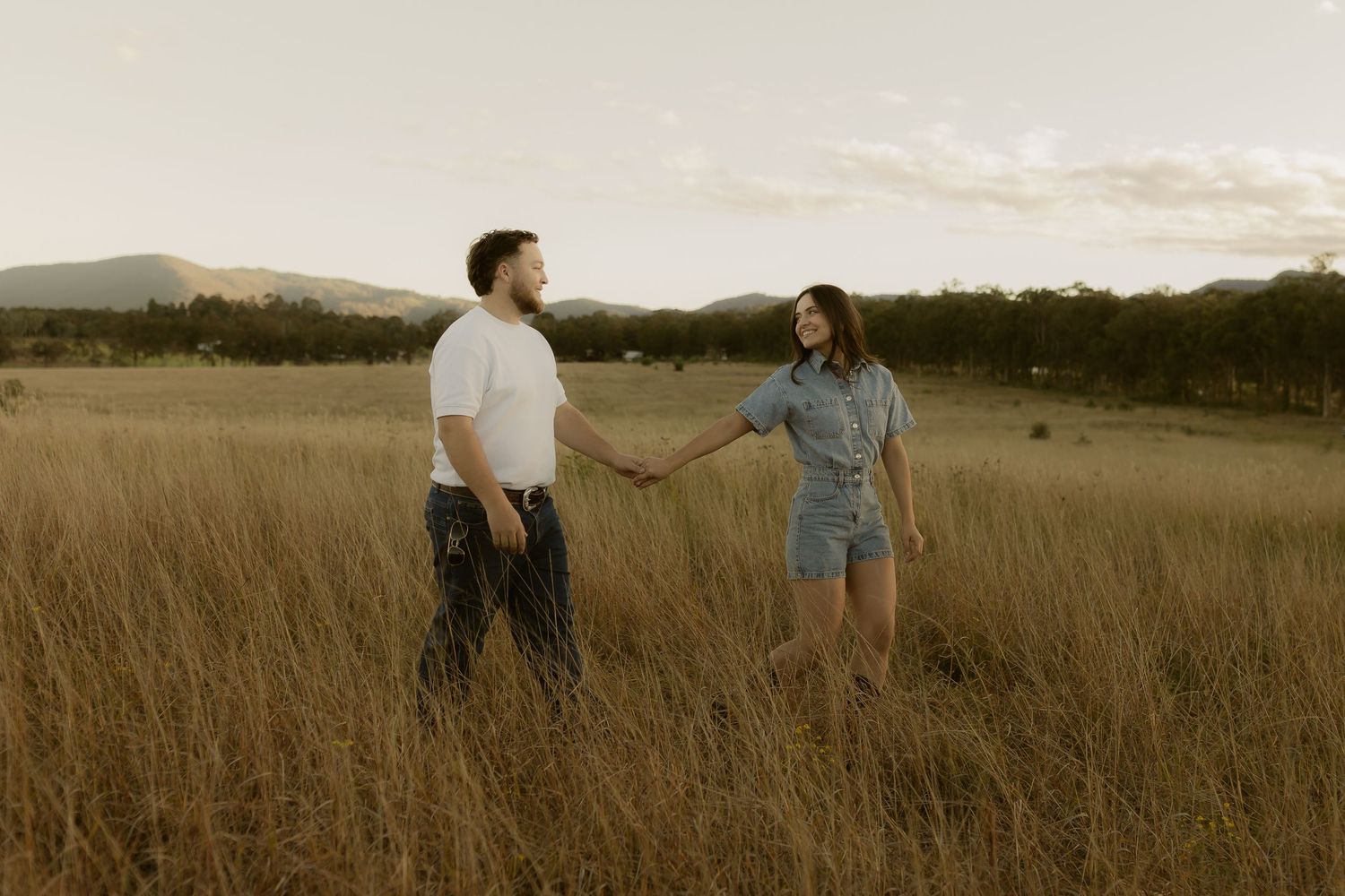 Couple holding hands facing each other in open golden grass field with mountains in background.
