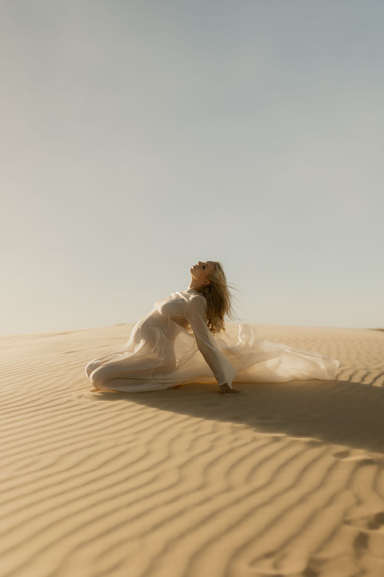 Woman in flowing white dress kneeling on golden sand dunes, hair blowing in wind against a soft blue sky.