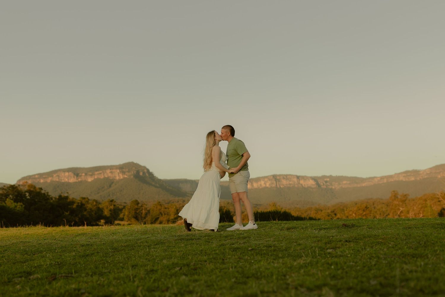 Couple sharing a romantic kiss at golden hour with stunning Blue Mountains backdrop on lush green lawn.