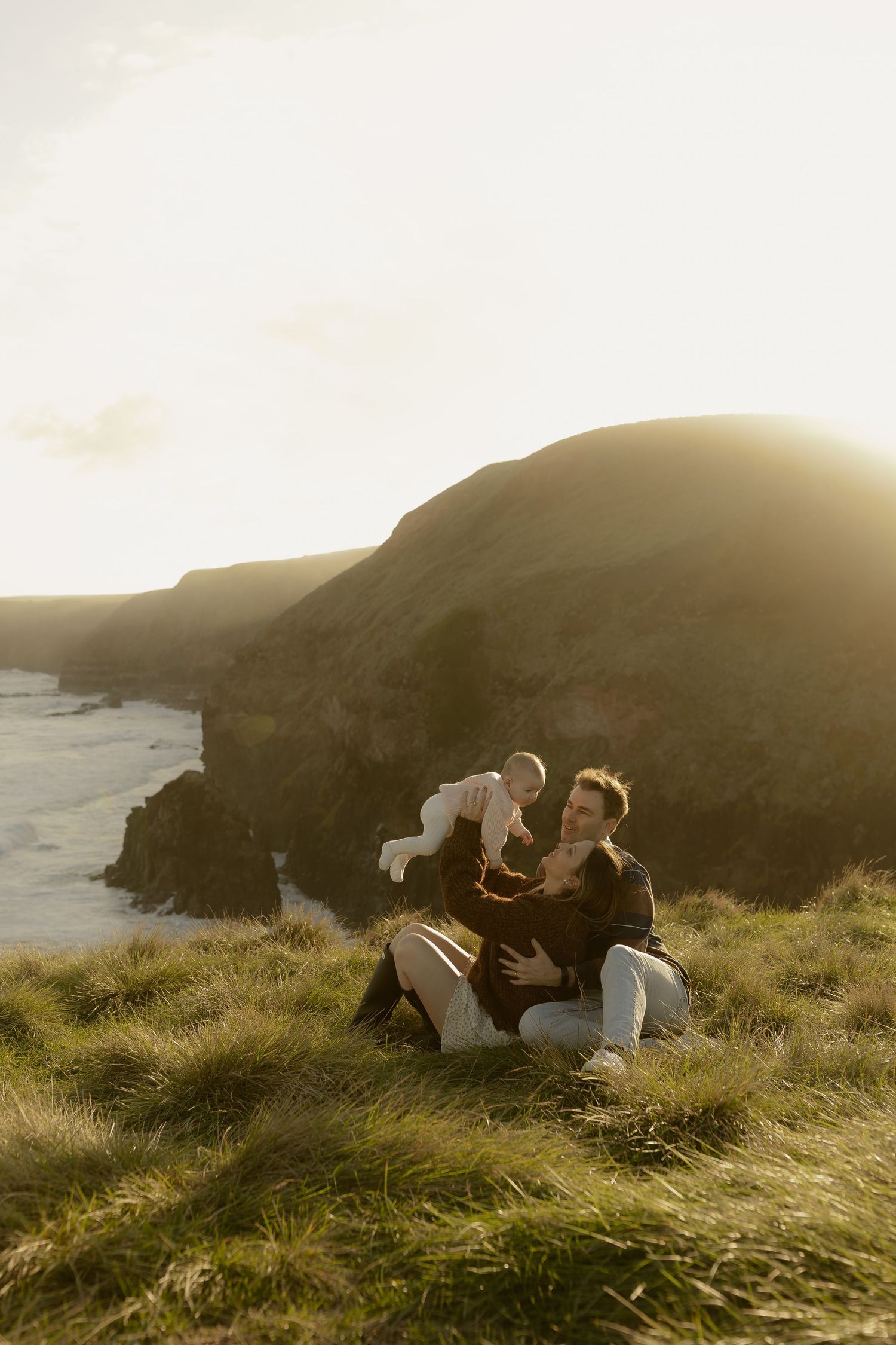 A man playfully lifts a baby while sitting on a grassy coastal cliff at sunset.