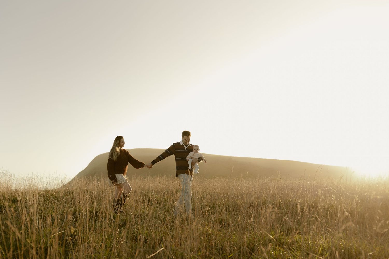 Couple holding hands in golden sunset light in a grassy field with a hill in the background.