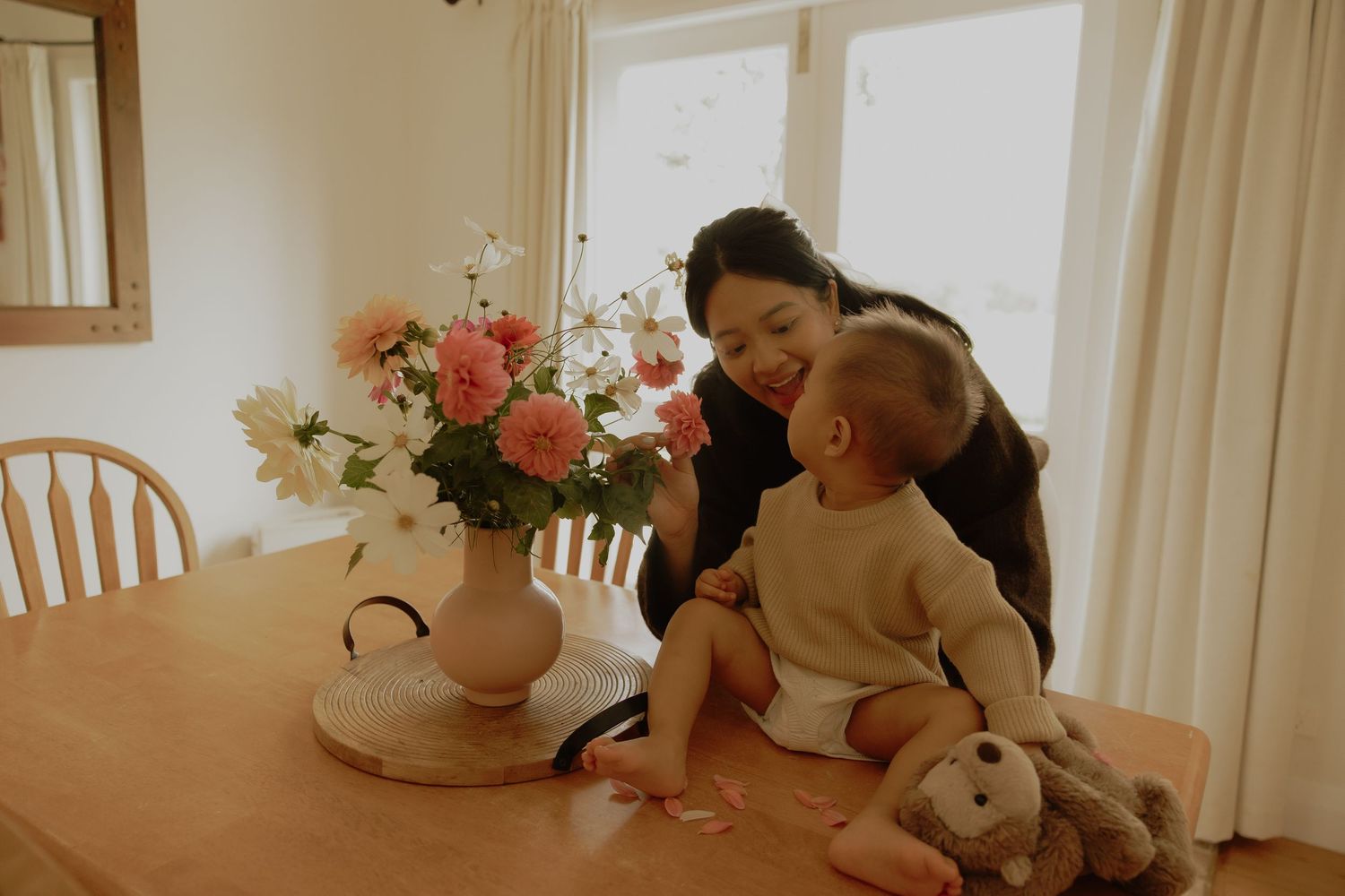 A mother smiles at her baby sitting on a wooden dining table beside a pink floral arrangement in a bright room.