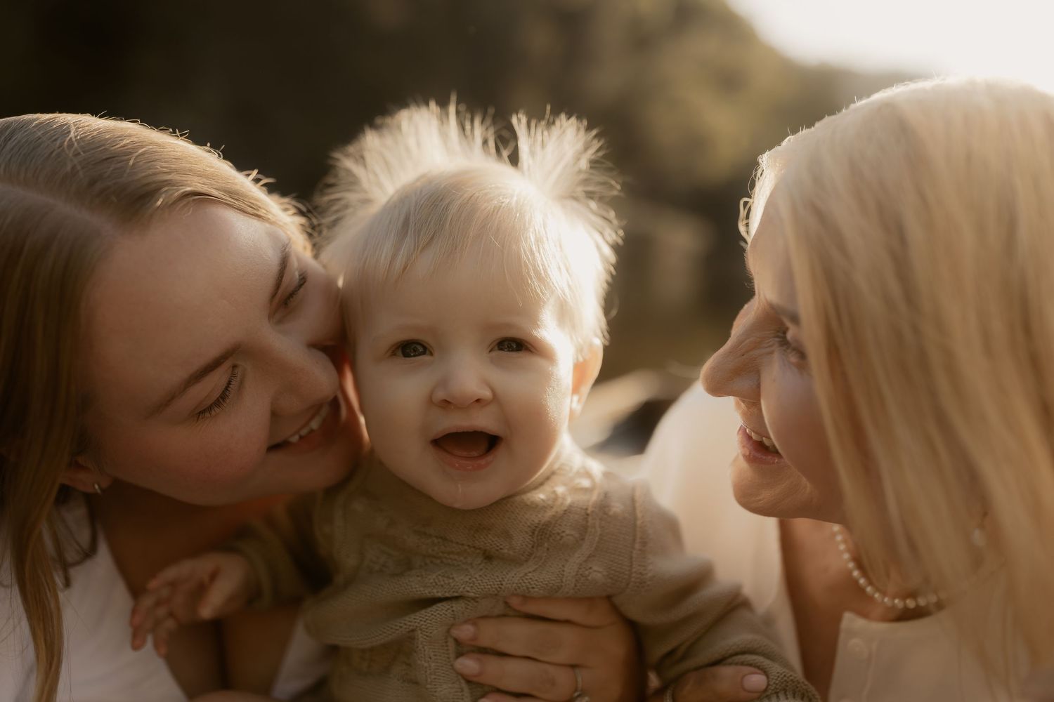 Two women lovingly hold a happy laughing baby outdoors in warm golden sunlight.