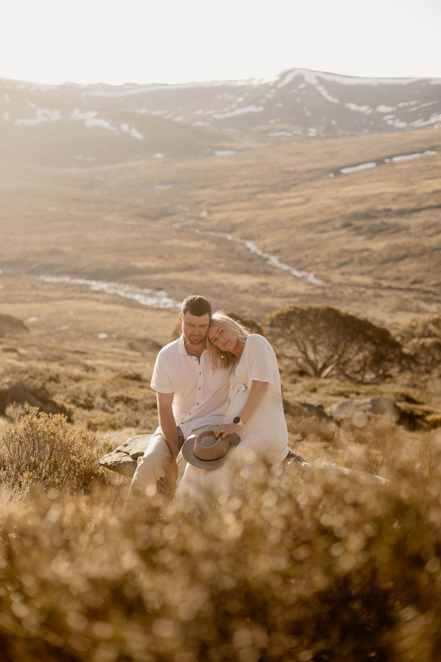 A couple embraces on a mountainside during golden hour with rolling hills and valleys stretching into the distance.