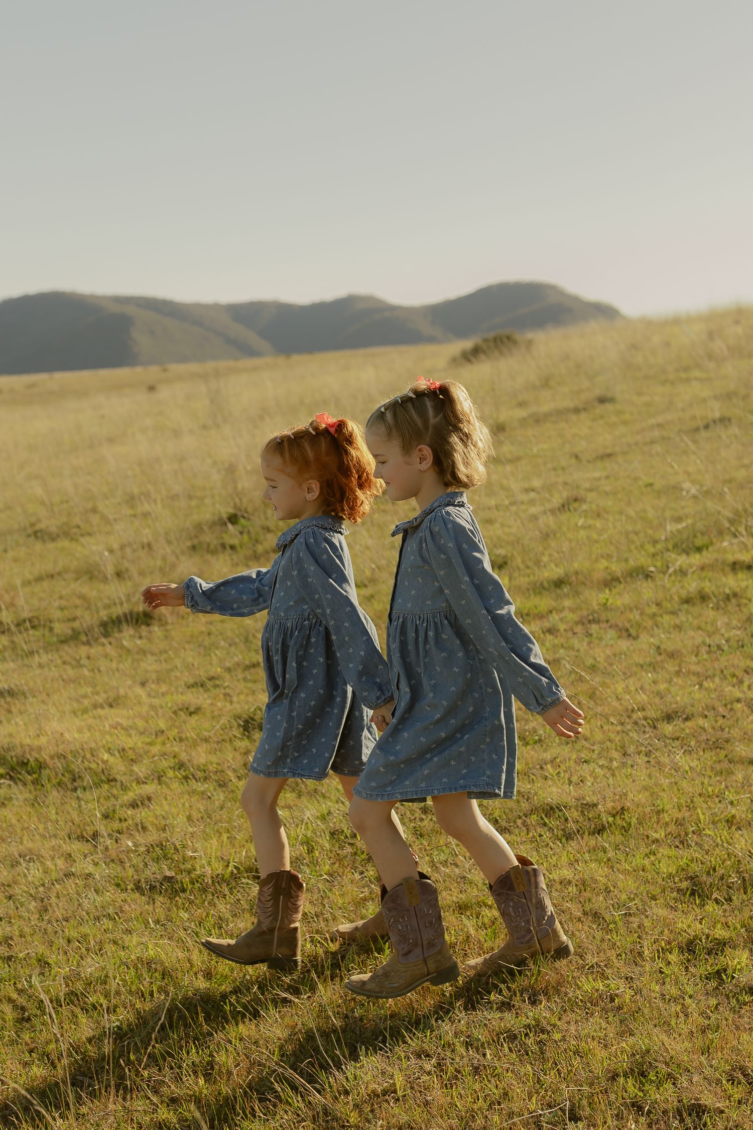 Two young girls in matching blue dresses and cowboy boots walk through a sunlit green meadow with rolling hills.