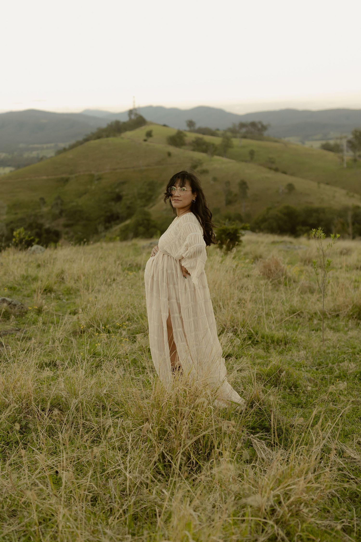 Pregnant woman in flowing white dress stands in green meadow with rolling hills backdrop.
