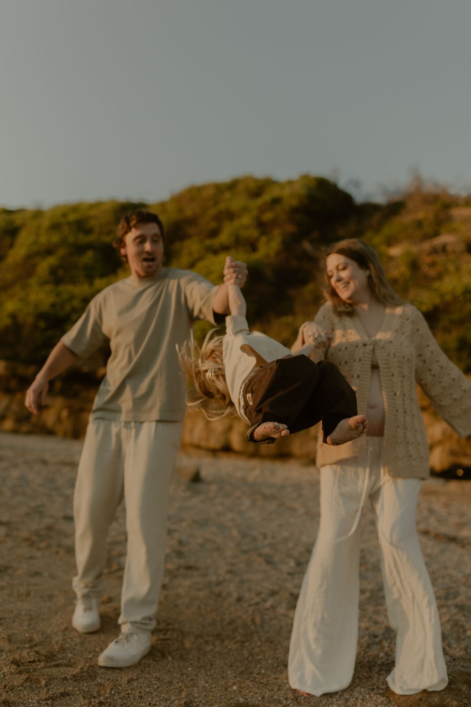 A joyful couple swings their child upside down on a sandy beach at golden hour, all dressed in neutral tones.