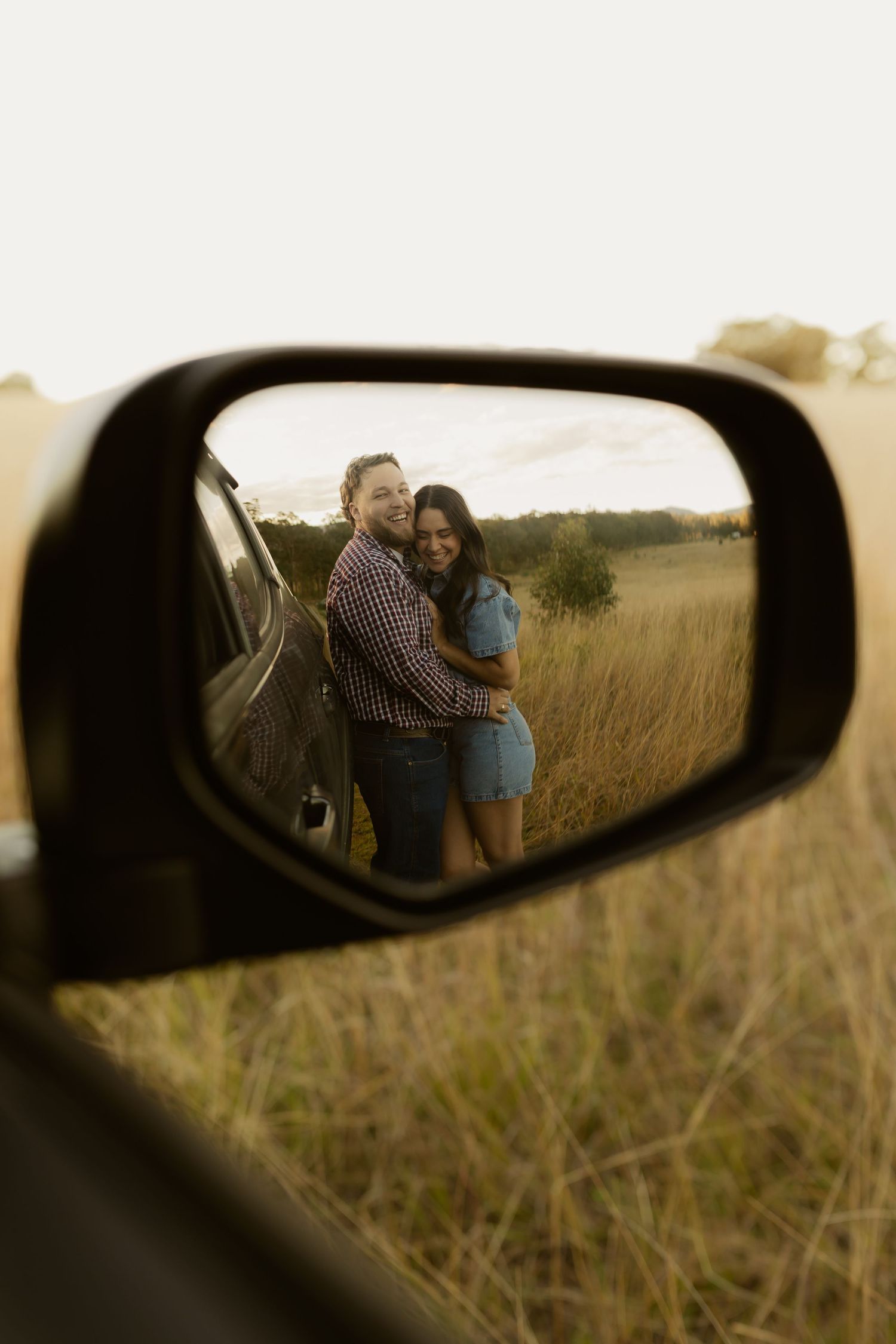 Couple embracing in golden field reflected in car side mirror during sunset road trip.
