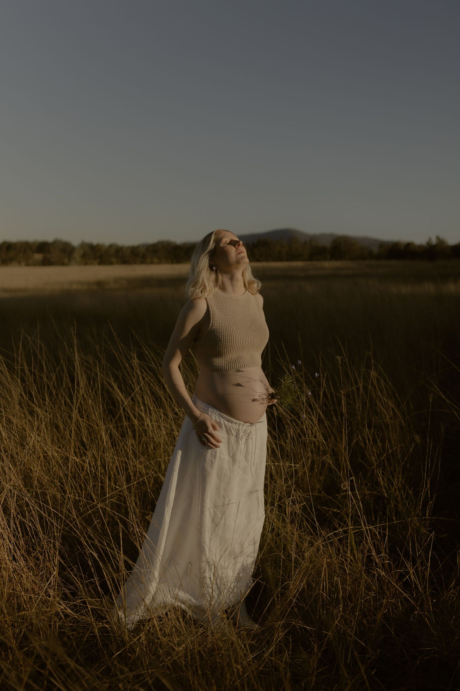 Blonde woman in beige top and white maxi skirt standing in golden grass field at dusk with mountains in background.