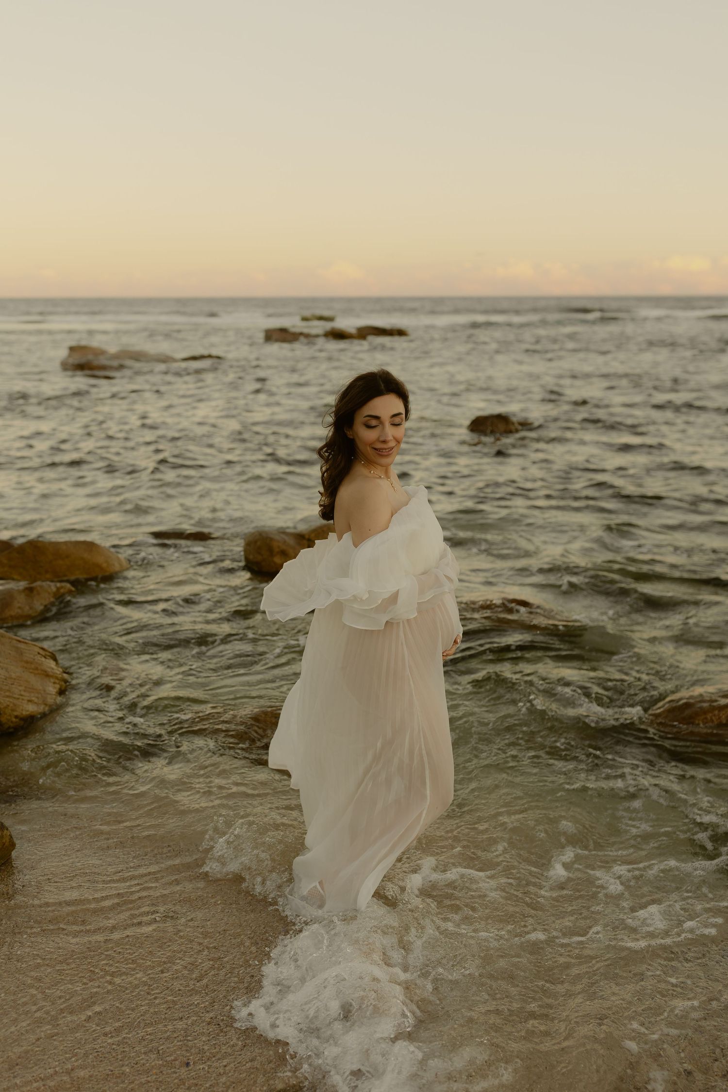 Smiling bride in flowing white dress wading through ocean waves at sunset.