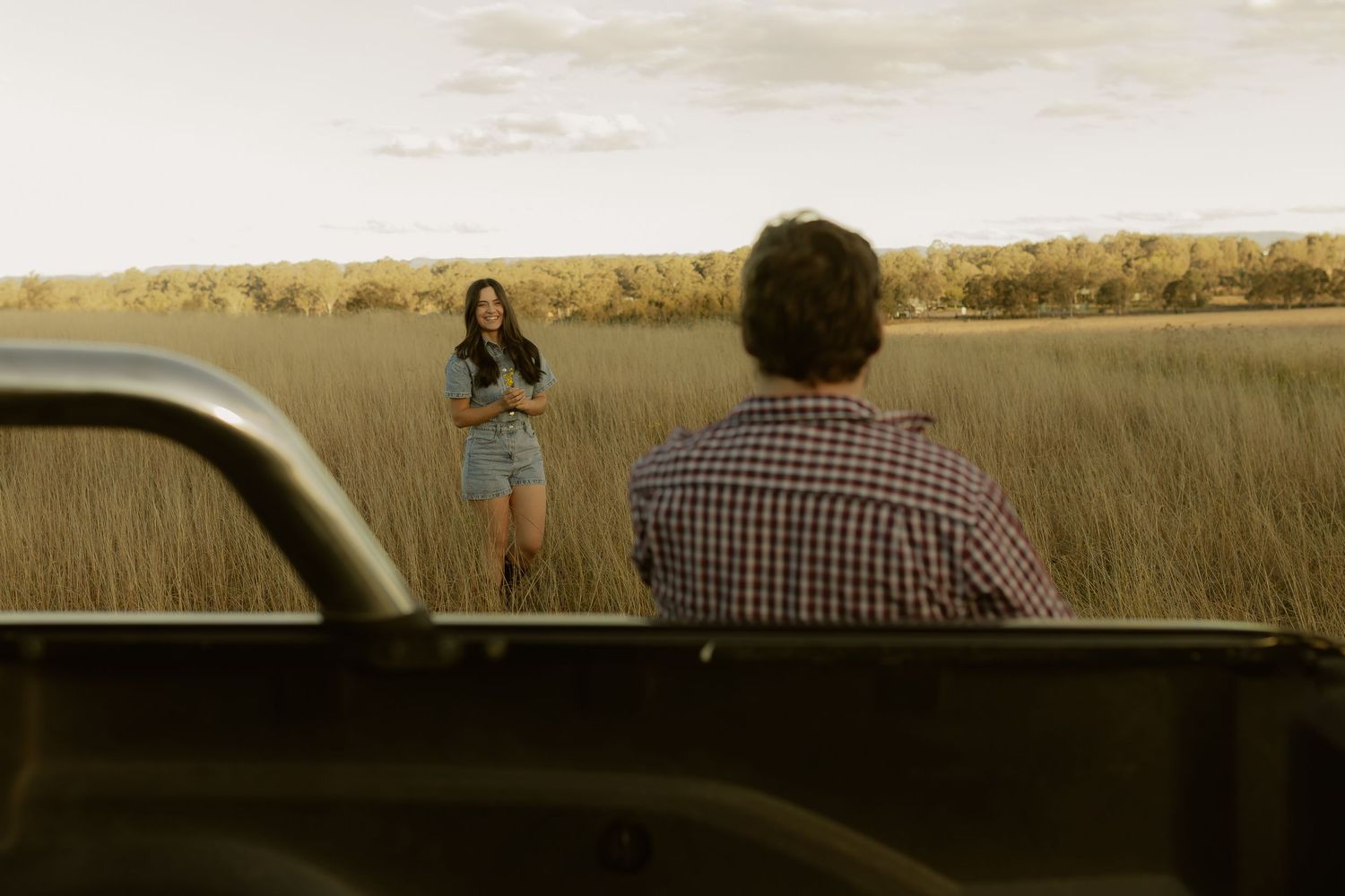 A woman stands in a golden field beside a truck as a man watches from the vehicle's bed.