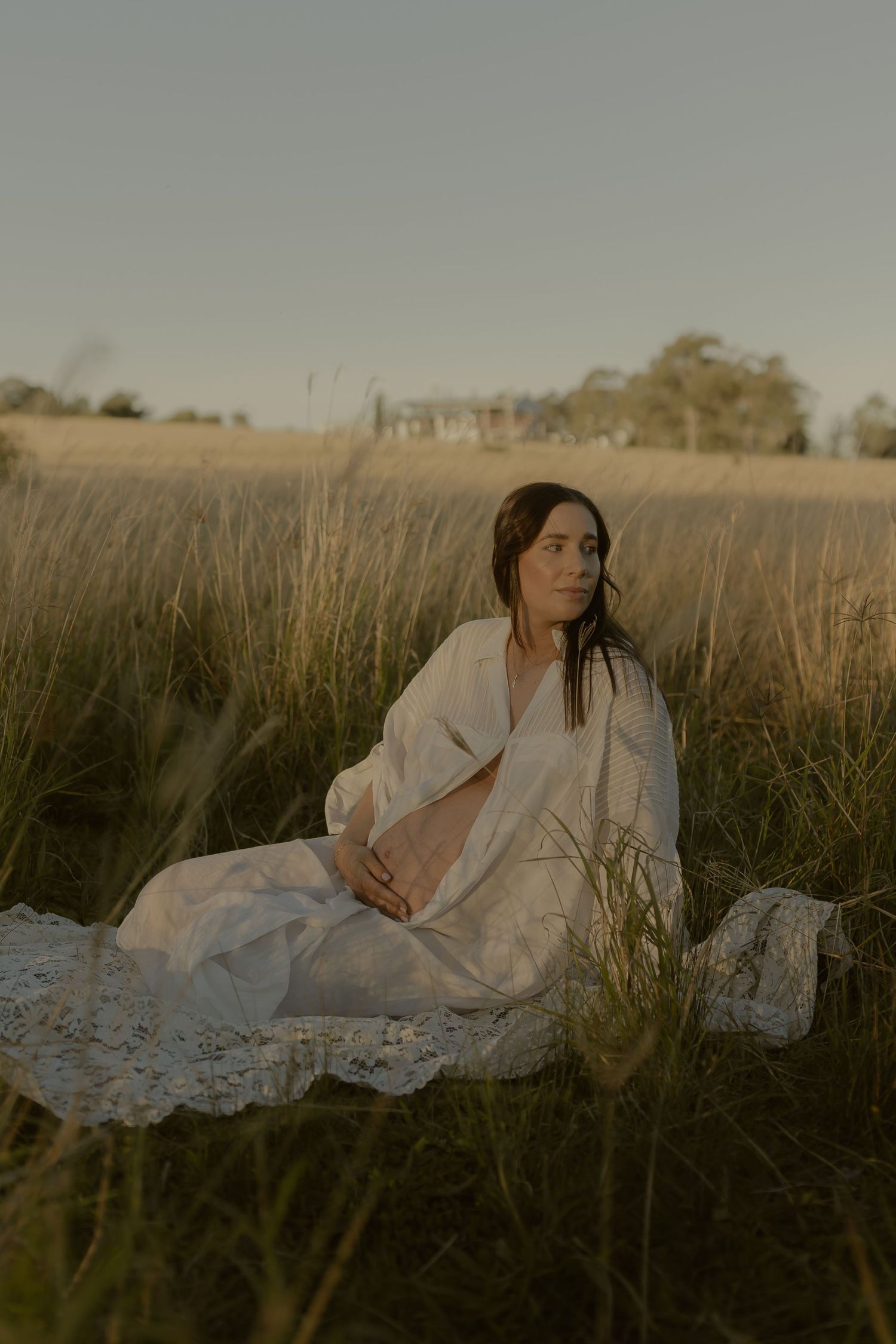 Pregnant woman in white robe sitting in tall golden grass field during warm sunset light.