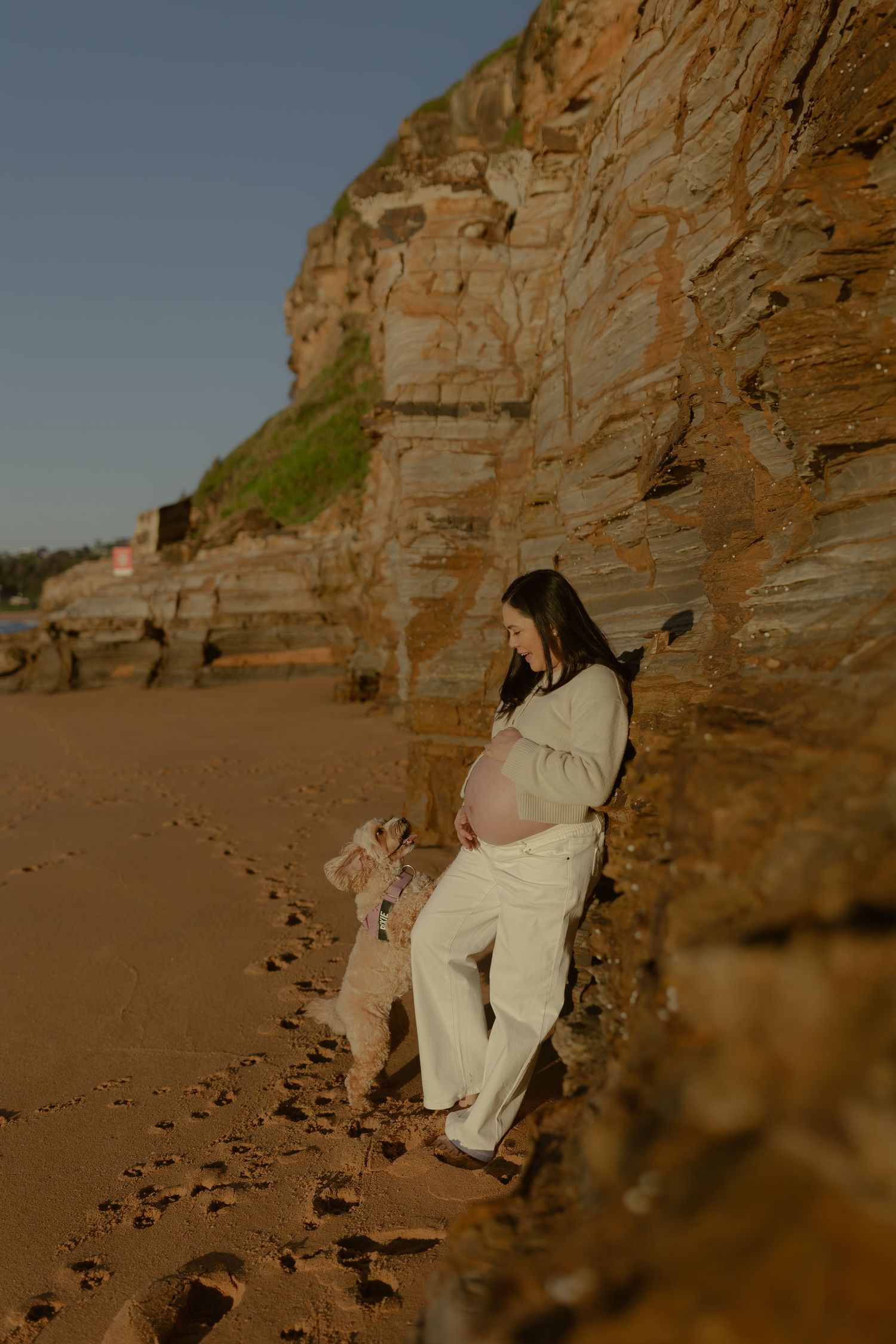 Pregnant woman in white outfit leaning against rocky cliff on sandy beach, tenderly cradling her baby bump.