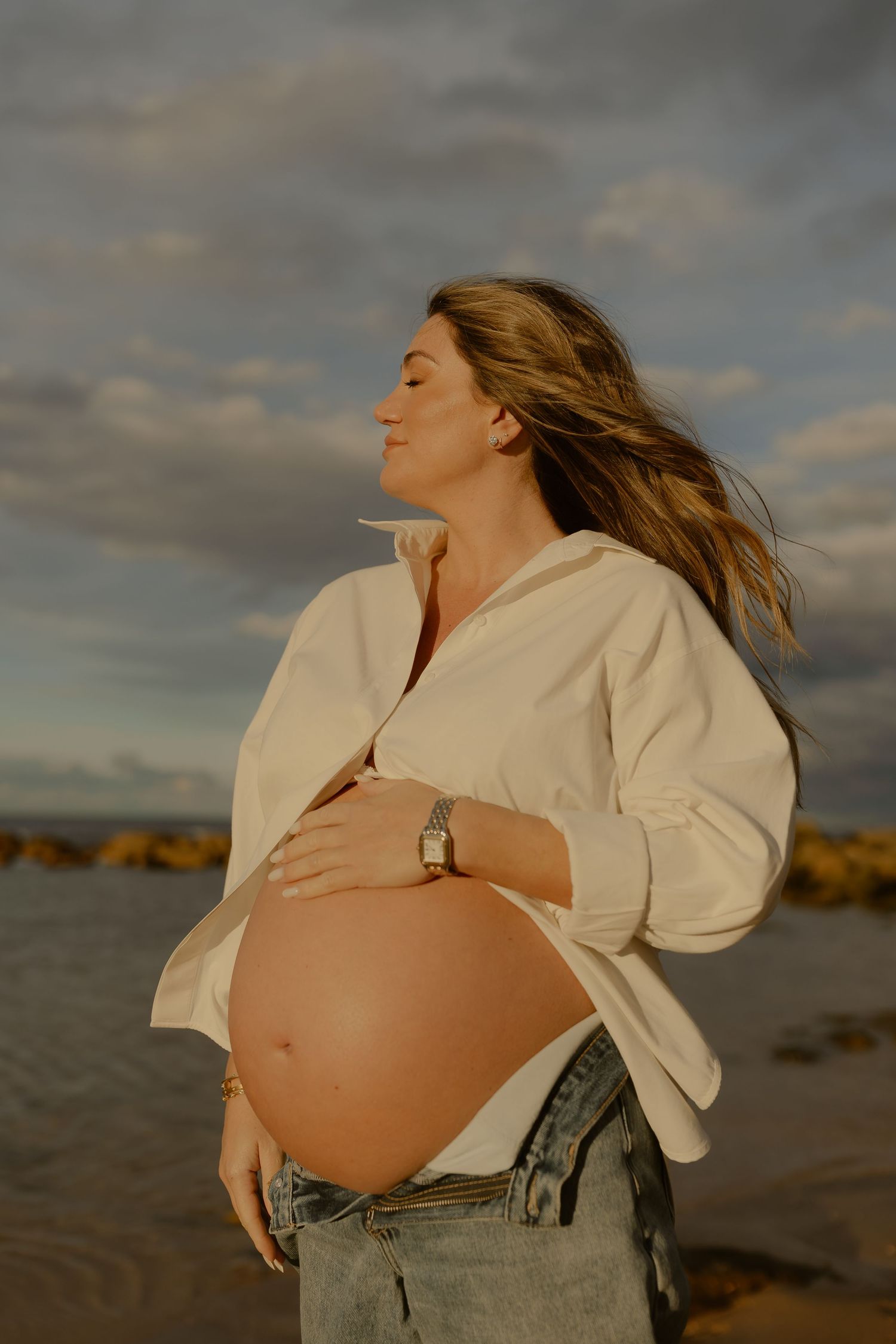 Pregnant woman in white shirt and jeans holds her bare belly on a beach at golden hour.