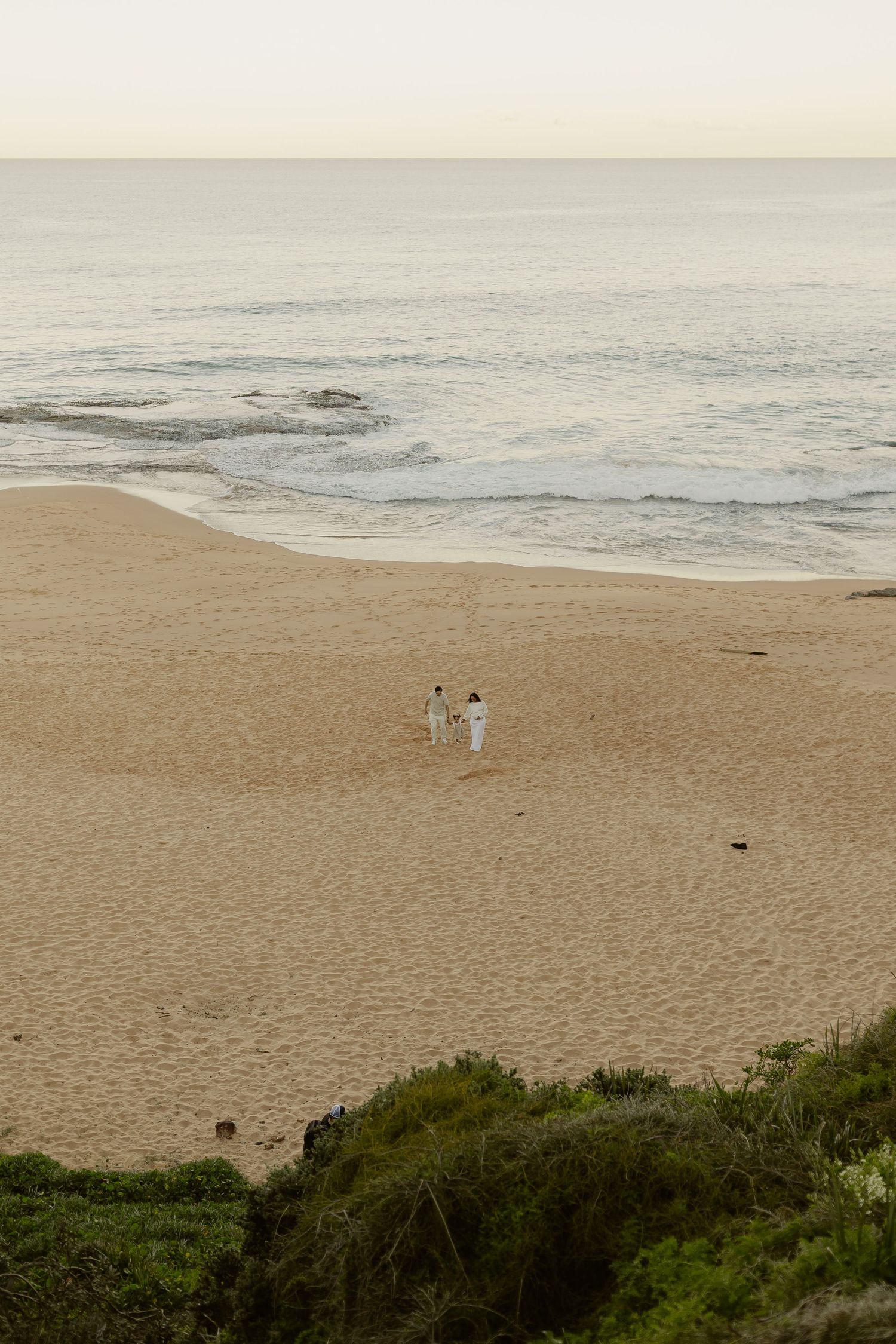 Couple in white walking hand-in-hand on a sandy beach at dusk with gentle ocean waves.