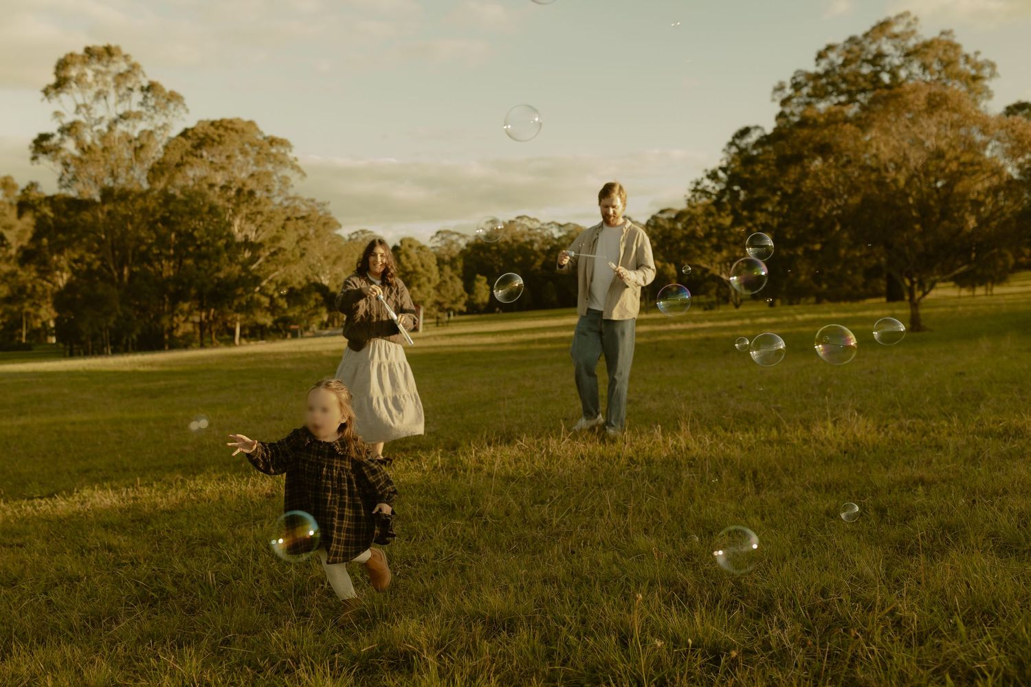 Family playing with bubbles in a sunny green park, parents watching toddler chase floating bubbles outdoors.