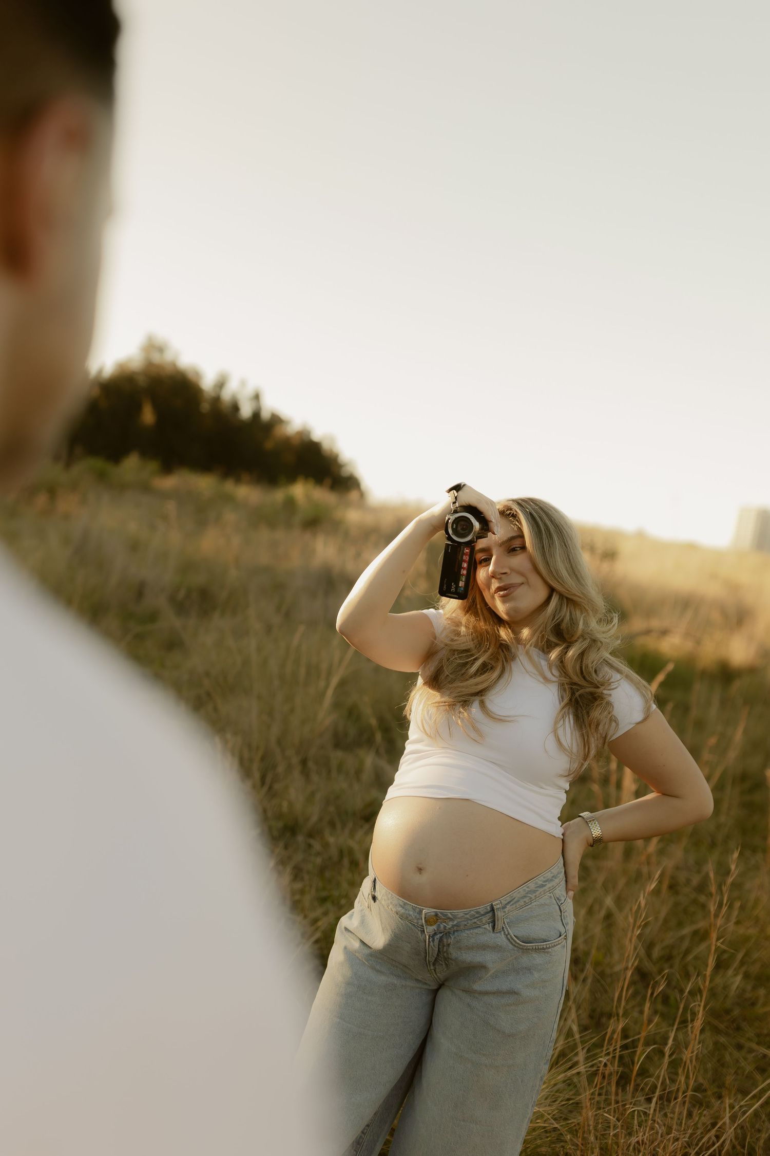 Pregnant woman in white crop top and jeans holds camera in golden hour field.